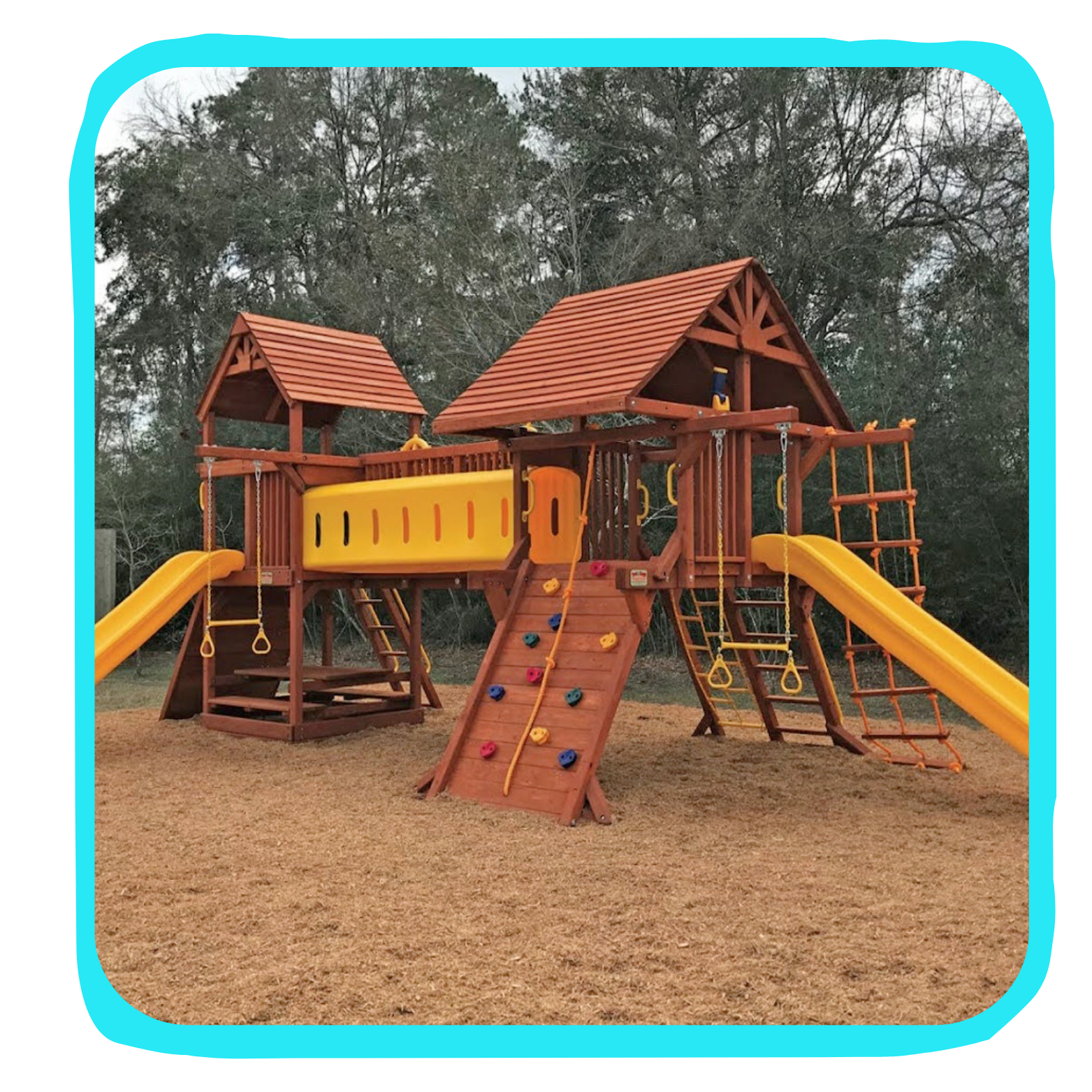 Large outdoor wooden playground with slides, climbing walls, and swings, set on a grassy field with trees in the background.
