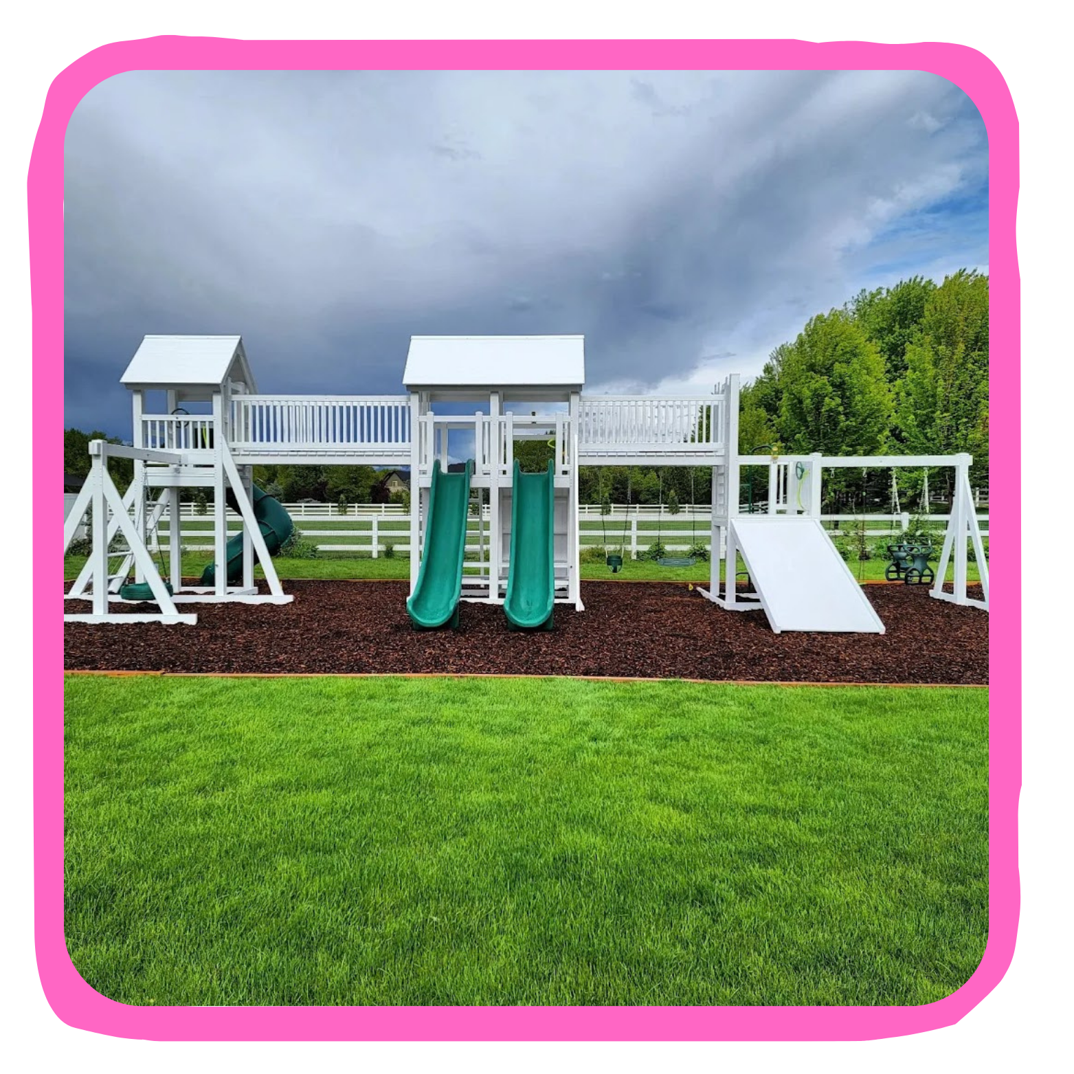 White playground structure with two slides, a climbing wall, swings, and a shaded platform, set on brown mulch with green grass and trees in the background.