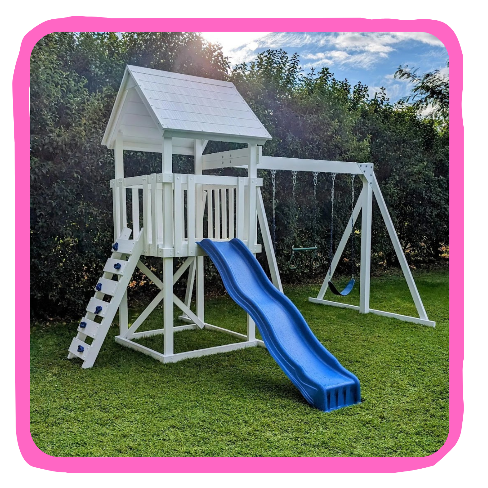 A white outdoor wooden playset with a slide, swing, and climbing wall, surrounded by green grass and bushes, under a partly cloudy sky.