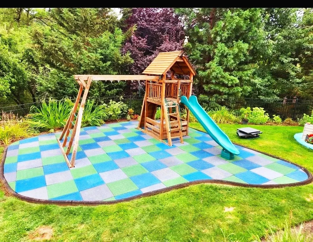 A backyard children's play ground with a wooden play structure featuring a slide, swing set, and a canopy, on a colorful rubberized safety surface surrounded by lush greenery and trees.