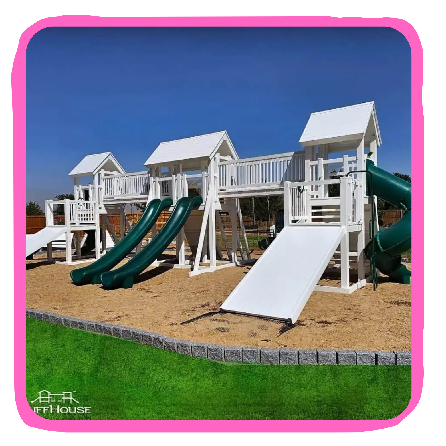 A white wooden playground structure with multiple slides and small house-like rooftops on a sandy ground with a green grassy border under a clear blue sky.