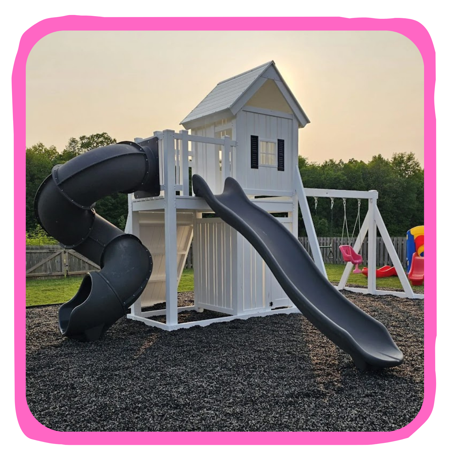 A backyard playground with a white wooden playhouse and two slides, one spiral and one straight, set on a gravel surface with a grassy area and trees in the background.