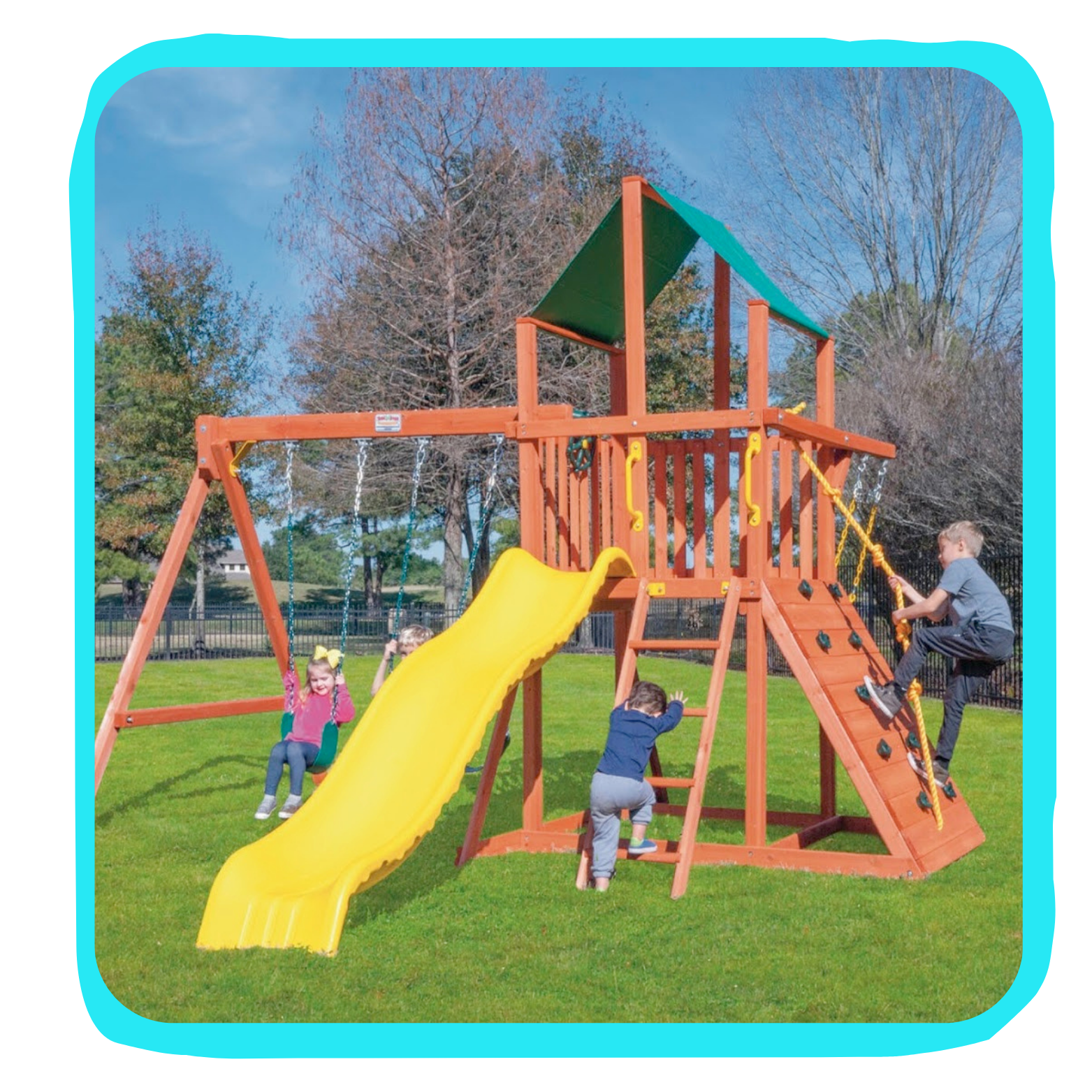 Children playing on a wooden playground with a yellow slide, swings, climbing wall, and a small roofed platform, on a grassy field.
