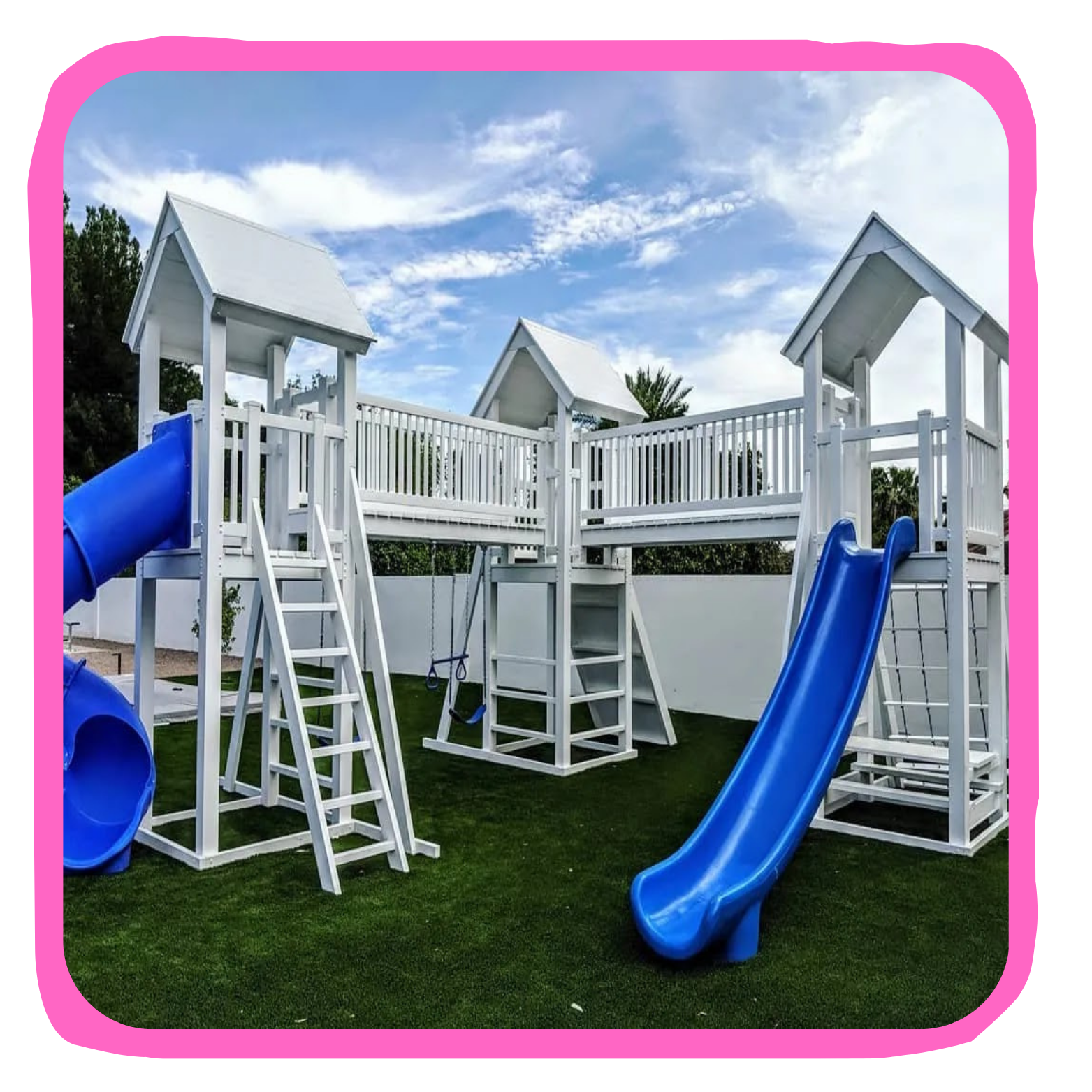 A white wooden playground structure with multiple levels, blue slides, and a small swing set on a well-maintained lawn under a cloudy sky.