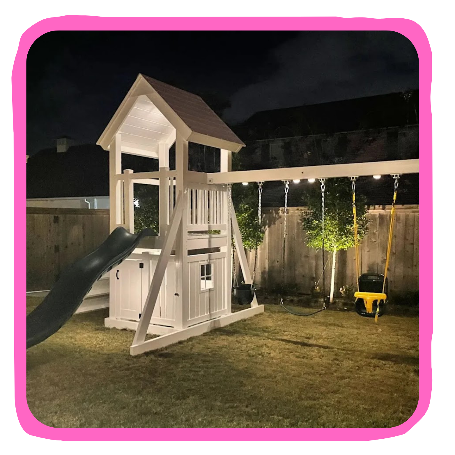 Nighttime backyard with a white wooden playset featuring a slide, swings, and a yellow toddler seat, surrounded by a wooden fence and small trees illuminated by outdoor lighting.