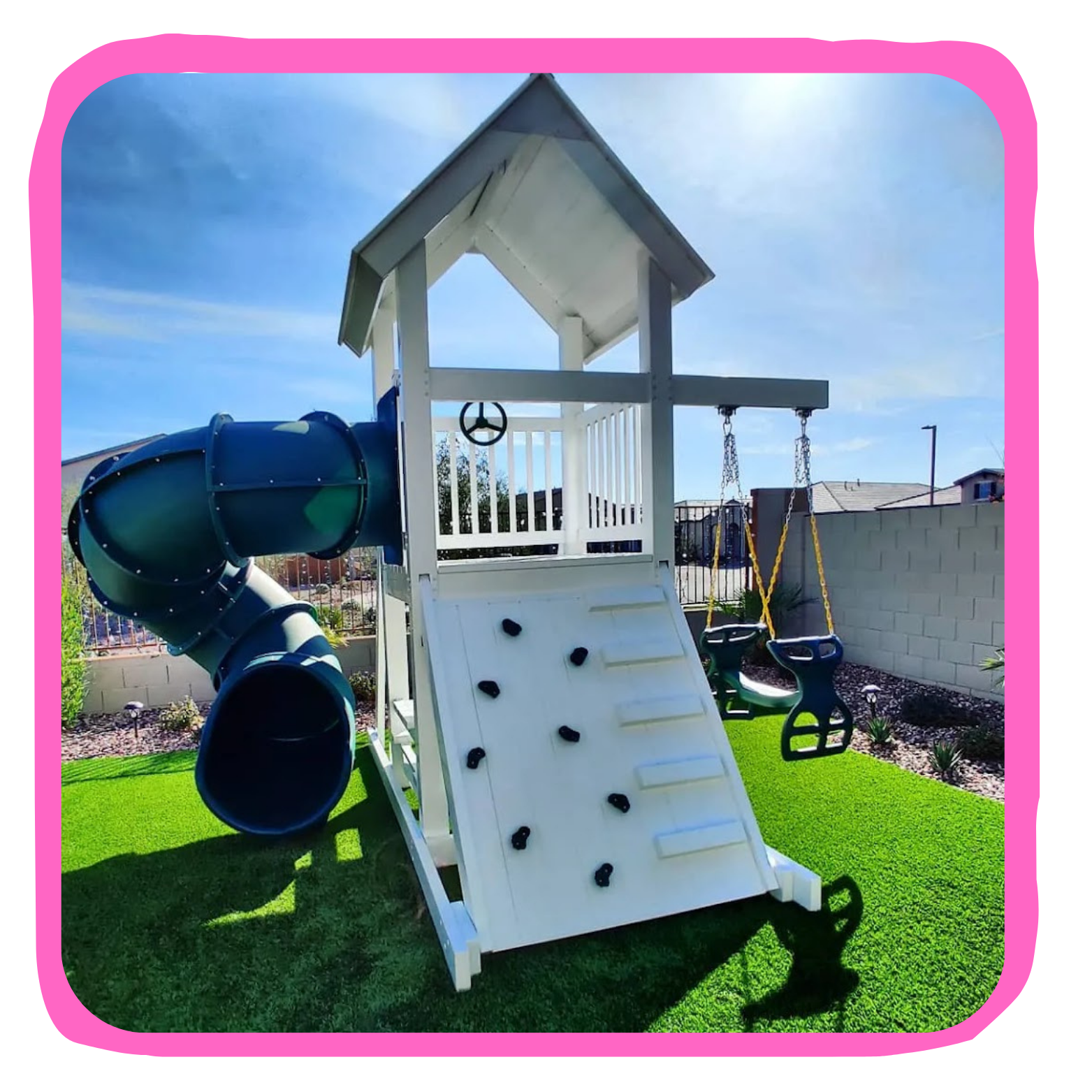 Children's outdoor play structure with a blue tube slide, a climbing wall, a small house with a roof, and two swings, set on a grassy yard with a brick wall and neighboring houses in the background.