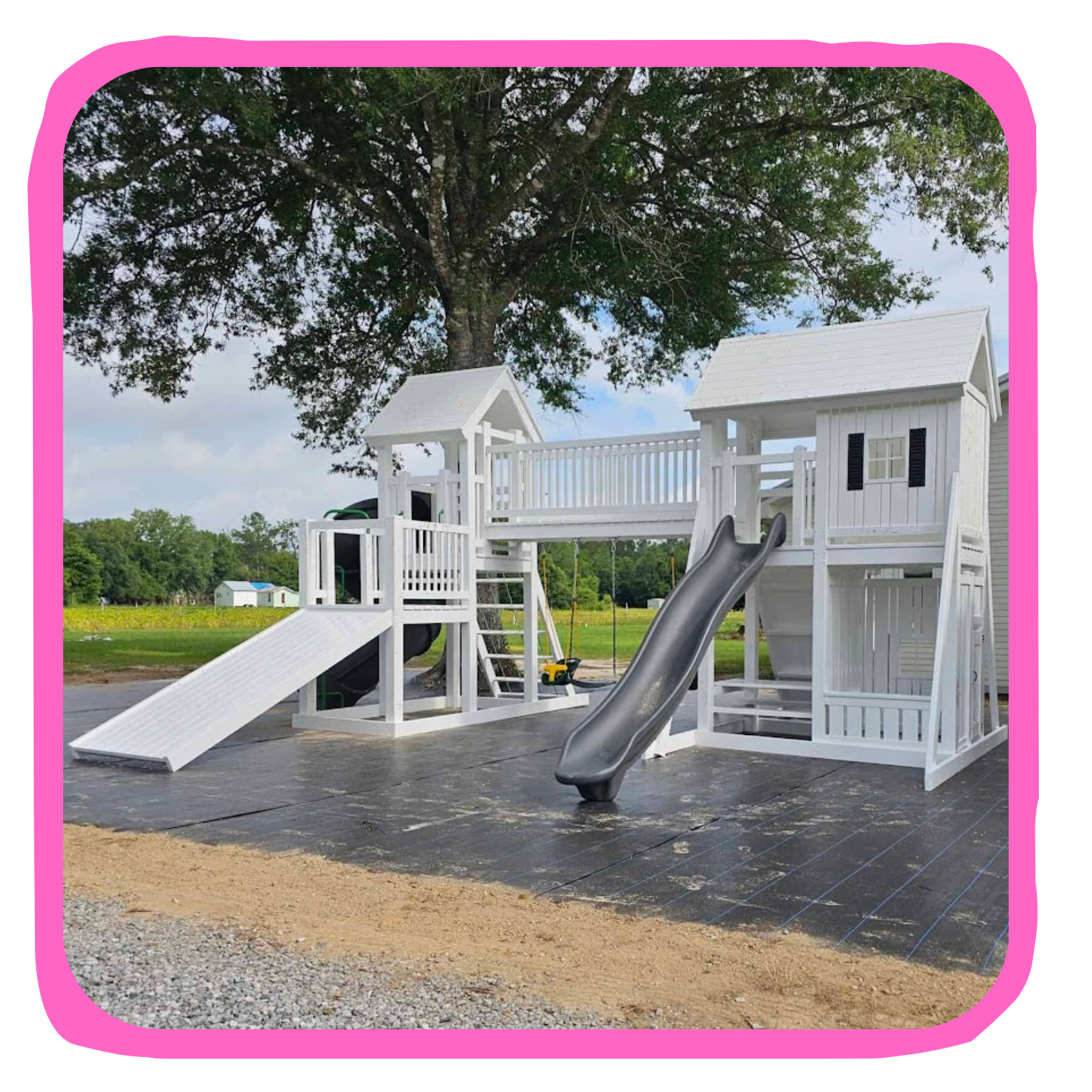 White wooden playhouse with slides and swings under a large tree on a black rubber surface.