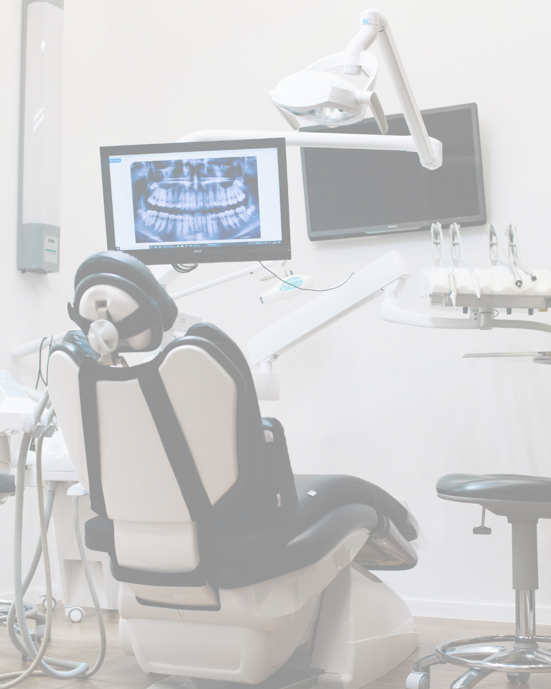 Dental clinic examination room with a dental chair, monitor displaying a panoramic dental X-ray, and dental equipment.