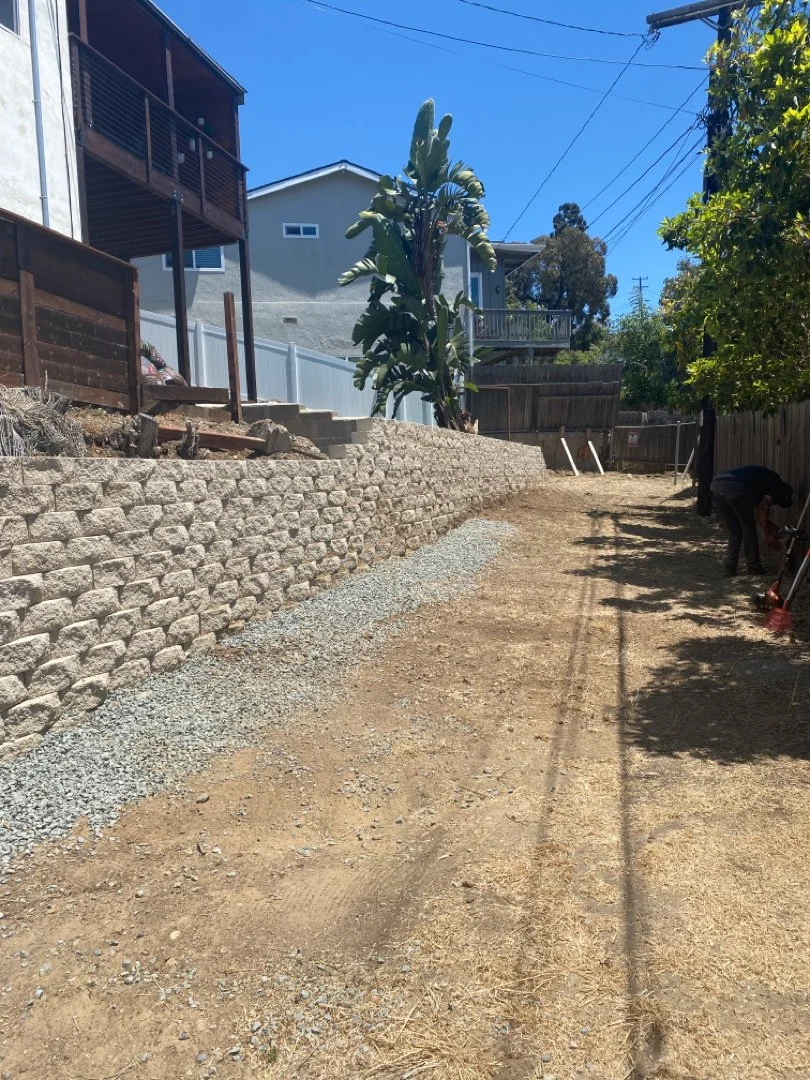 A backyard with a partially completed stone retaining wall and a dirt path. There are trees, a house, and utility wires in the background, with a person working near the right side.