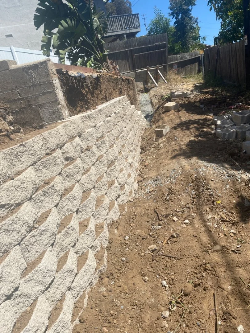 Construction work with a new stone retaining wall on the left side of the dirt pathway, and building materials scattered along the pathway, in a backyard with trees, wooden fences, and a deck in the background.