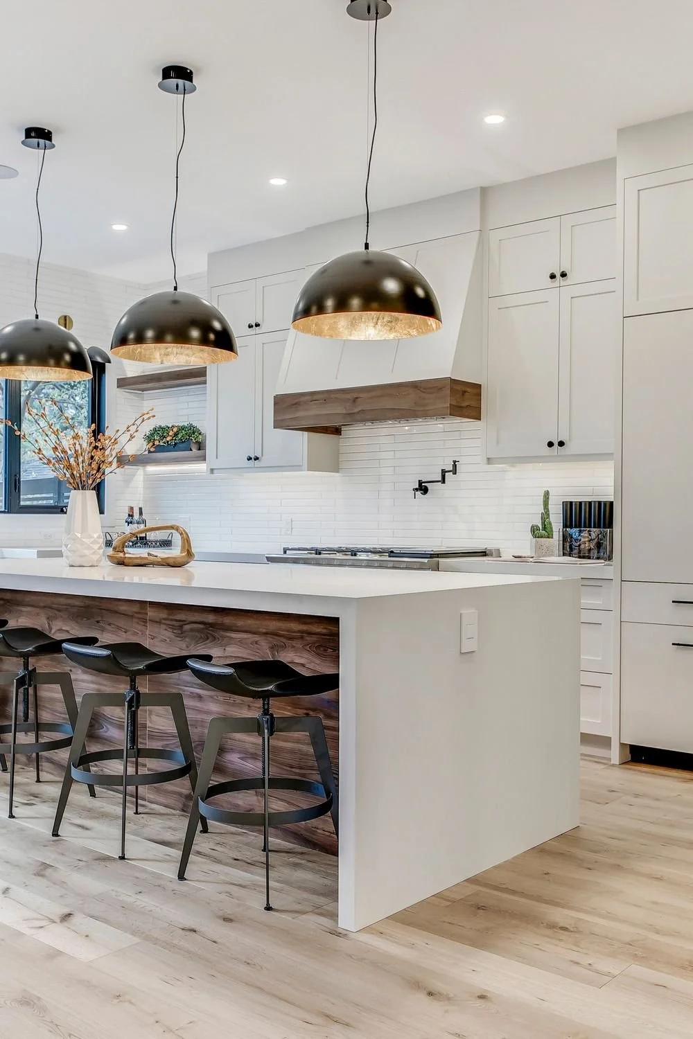 Modern kitchen with white cabinets, wooden accents, black pendant lights, and a kitchen island with three black chairs.
