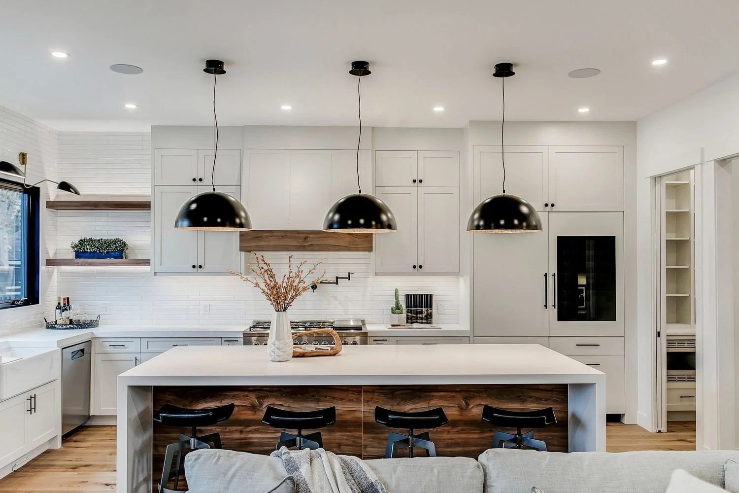 Modern white kitchen with black pendant lights over a large island, featuring a vase with dried flowers, surrounded by bar stools, with additional appliances and decor, hardwood floors.