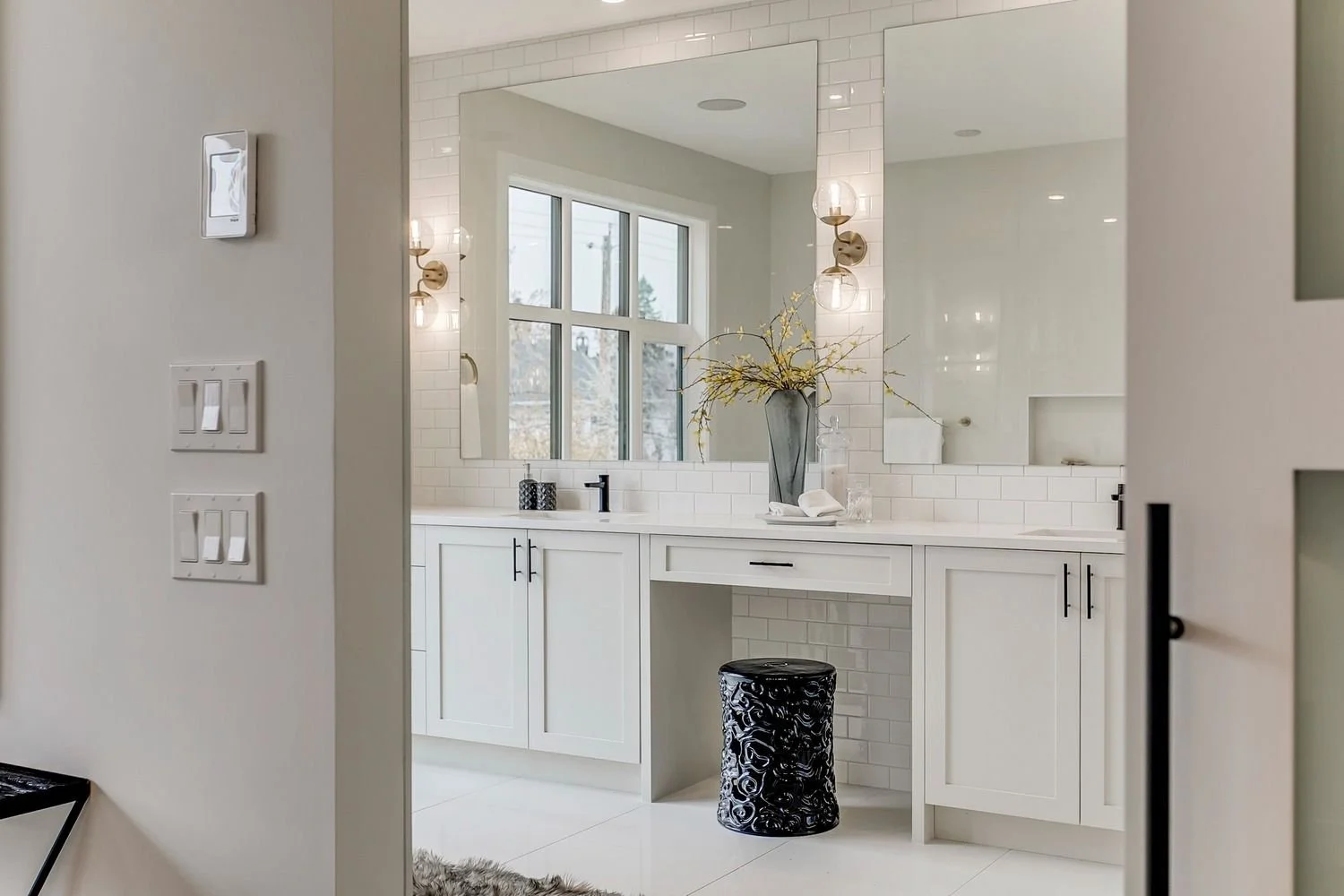 Modern bathroom with white vanity, large mirror, black fixtures, decorative black stool, and large window with natural light, white subway tile backsplash, and minimal decor.