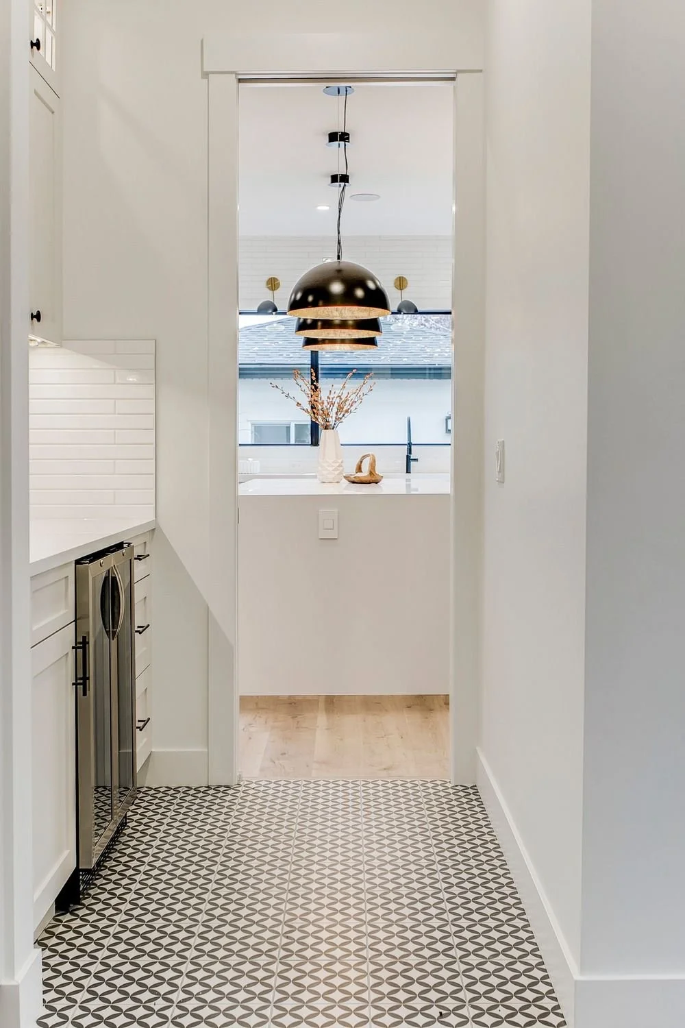 View from a small kitchen hallway looking into a bright room with a pendant light fixture, a white vase with dried branches, and a wooden tray on the counter.