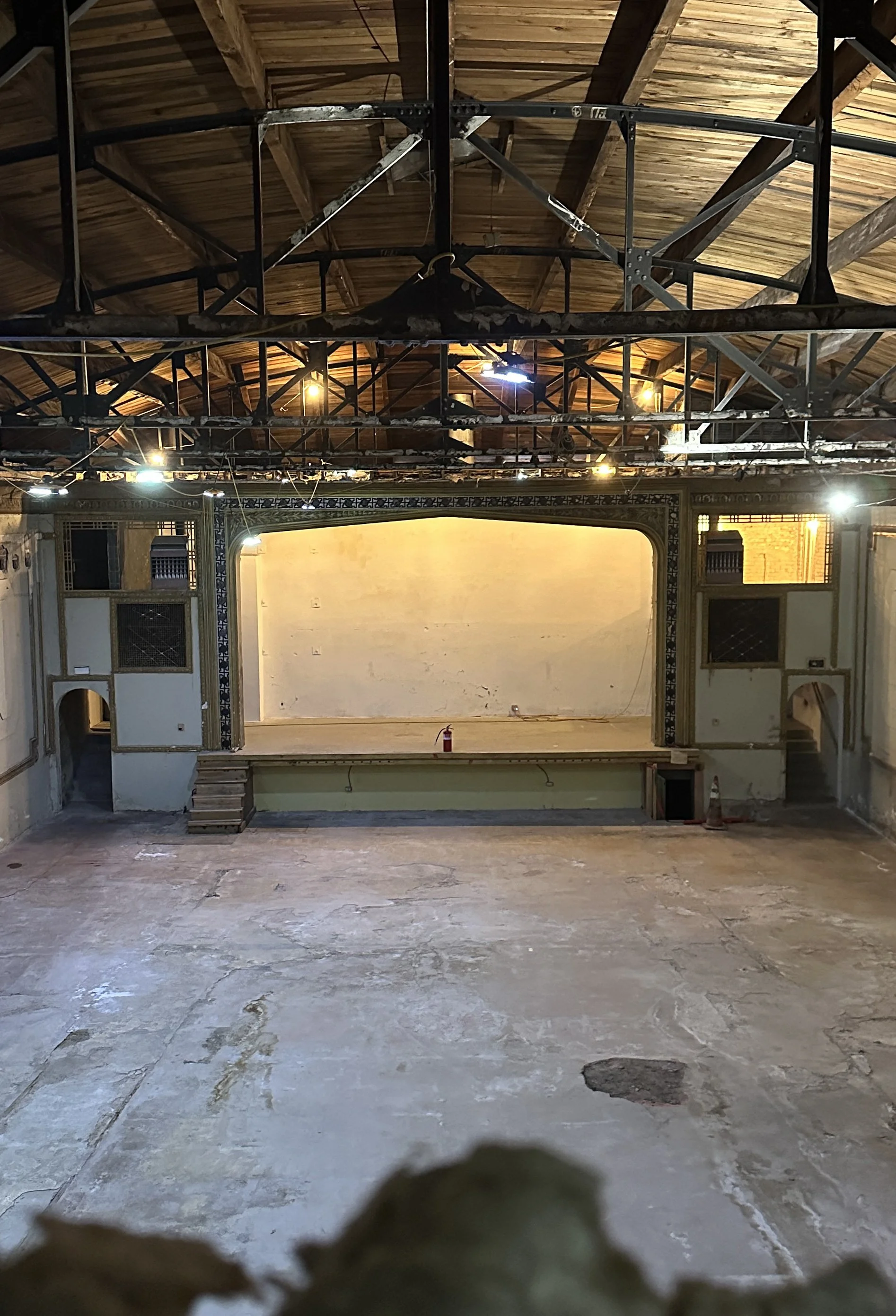An empty, historic theater with a worn-out floor, an elevated stage with a yellowed wall backdrop, and decorative architectural details around the stage area. The ceiling shows exposed wooden beams and metal support structures, all in need of repair.