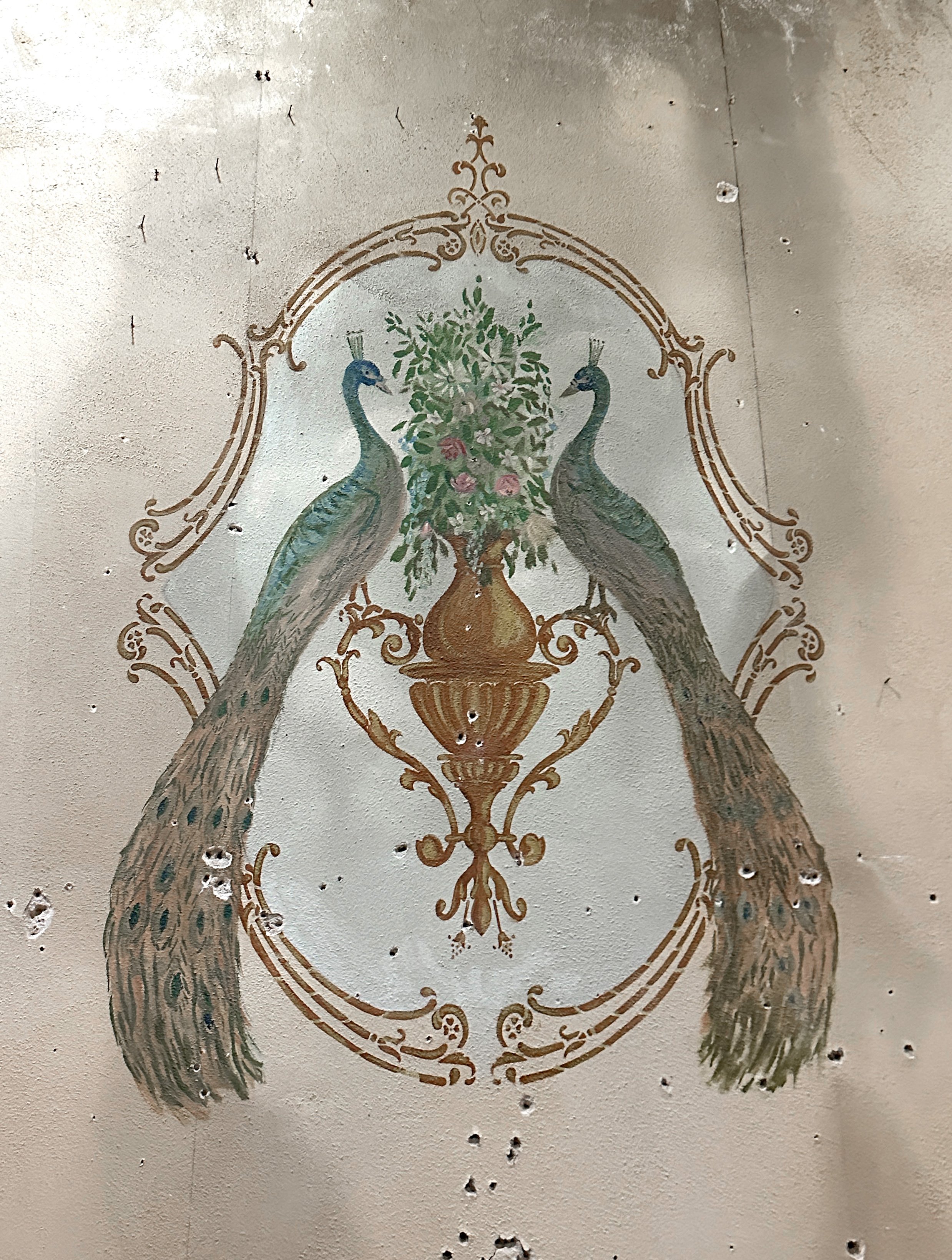 An early 20th century  wall mural of two peacocks standing on either side of a vase with flowers and green foliage, enclosed in an ornate gold frame. The wall is somewhat damaged and crumbling, and the mural is not perfectly intact.