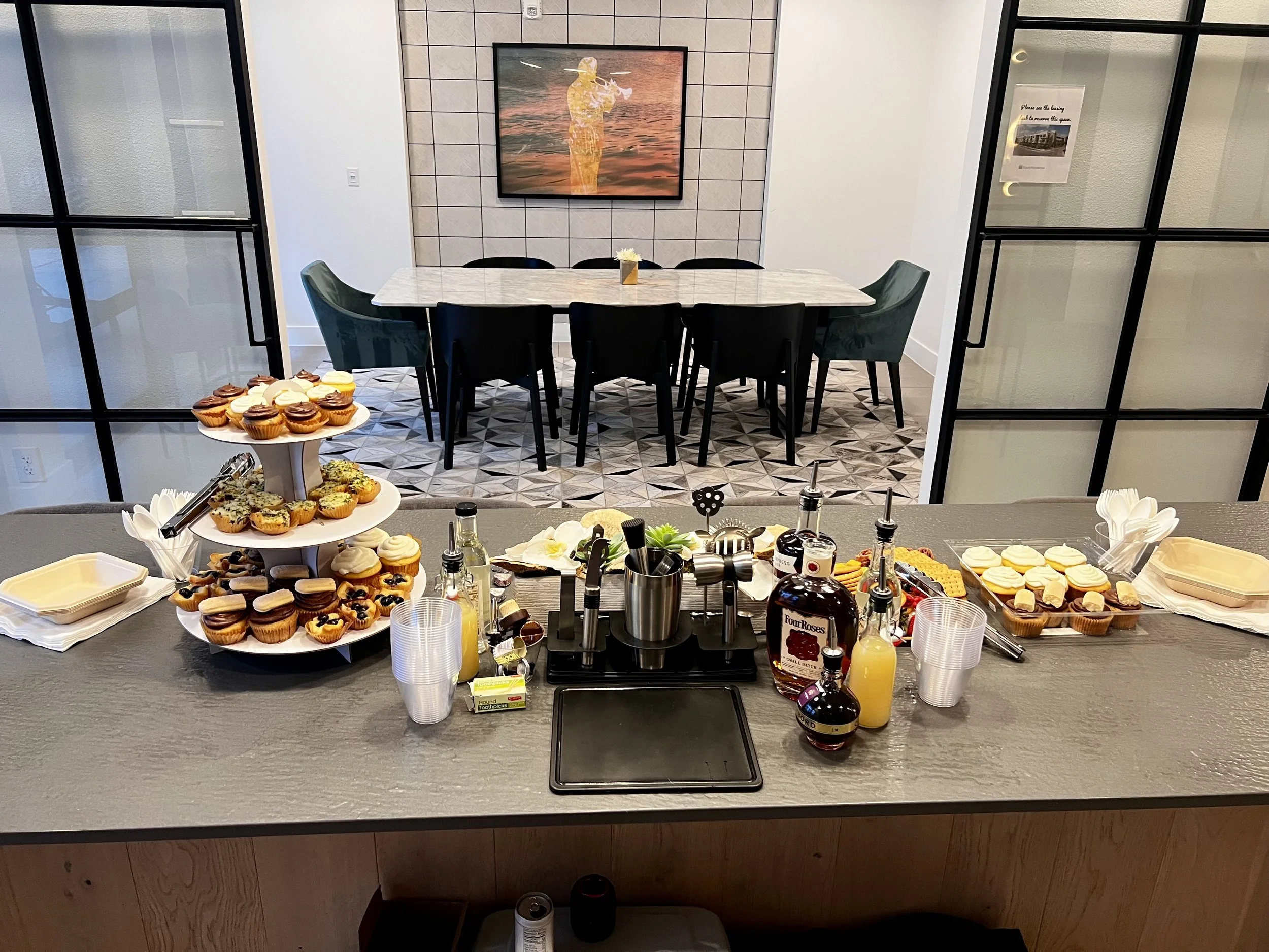 A breakfast or brunch buffet with cupcakes, muffins, and various breakfast items on a kitchen counter, with a dining room in the background featuring a table, chairs, and a framed artwork on the wall.