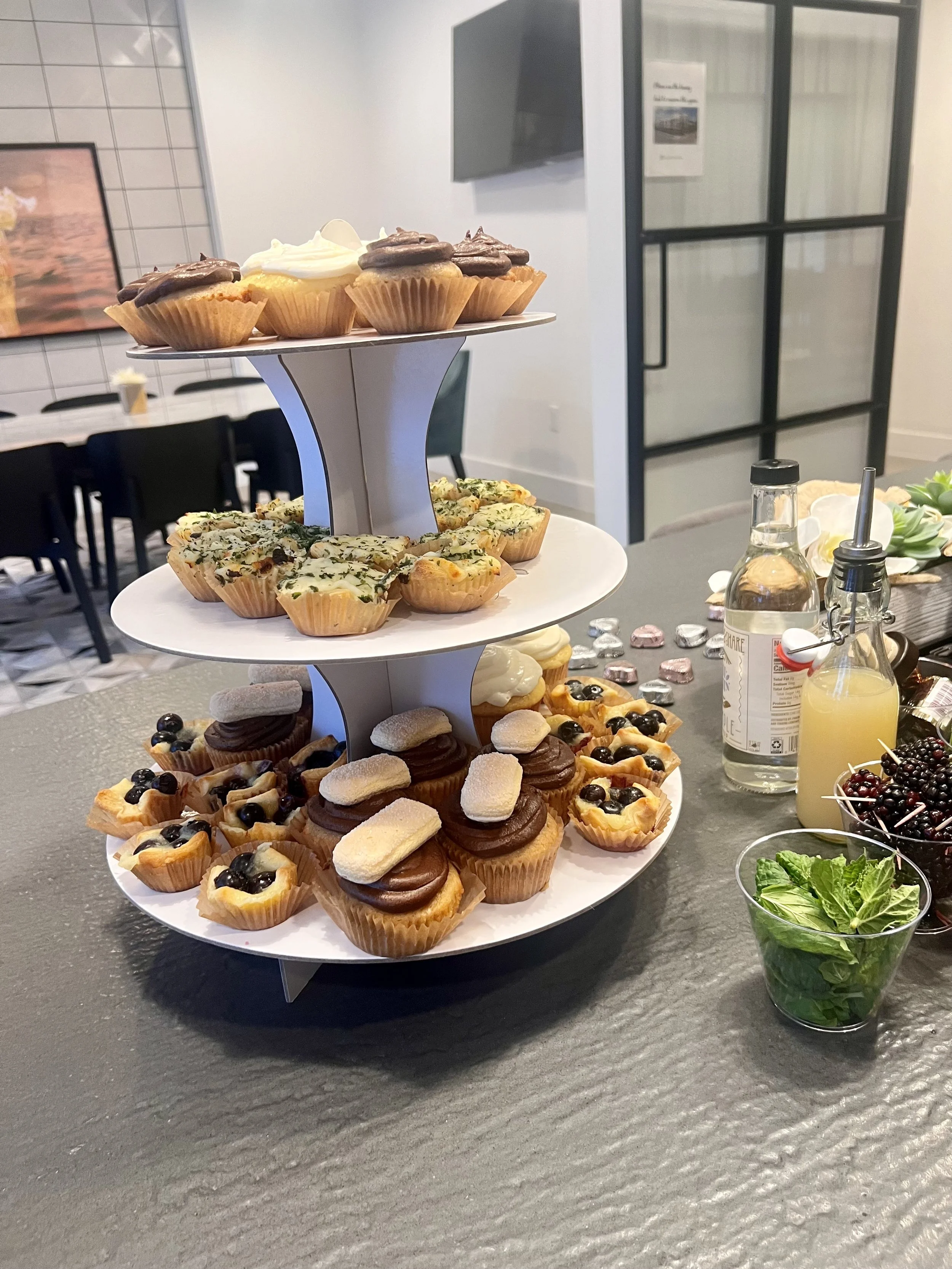 Tiered tray with assorted cupcakes, including chocolate, vanilla, and fruit-flavored varieties, arranged on a black table with garnishes like blackberries and mint nearby.