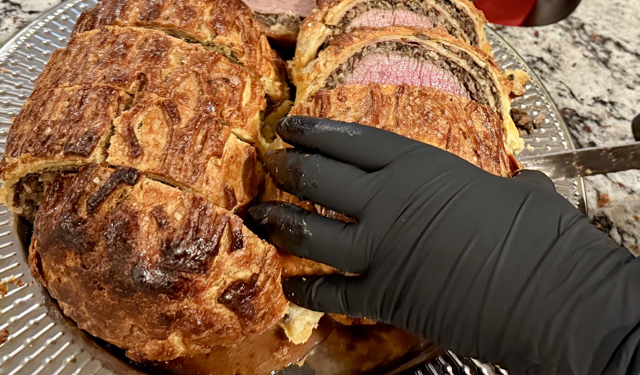 A person wearing a black glove carving a large beef Wellington on a silver platter, revealing the cooked beef and mushroom duxelles inside the pastry.