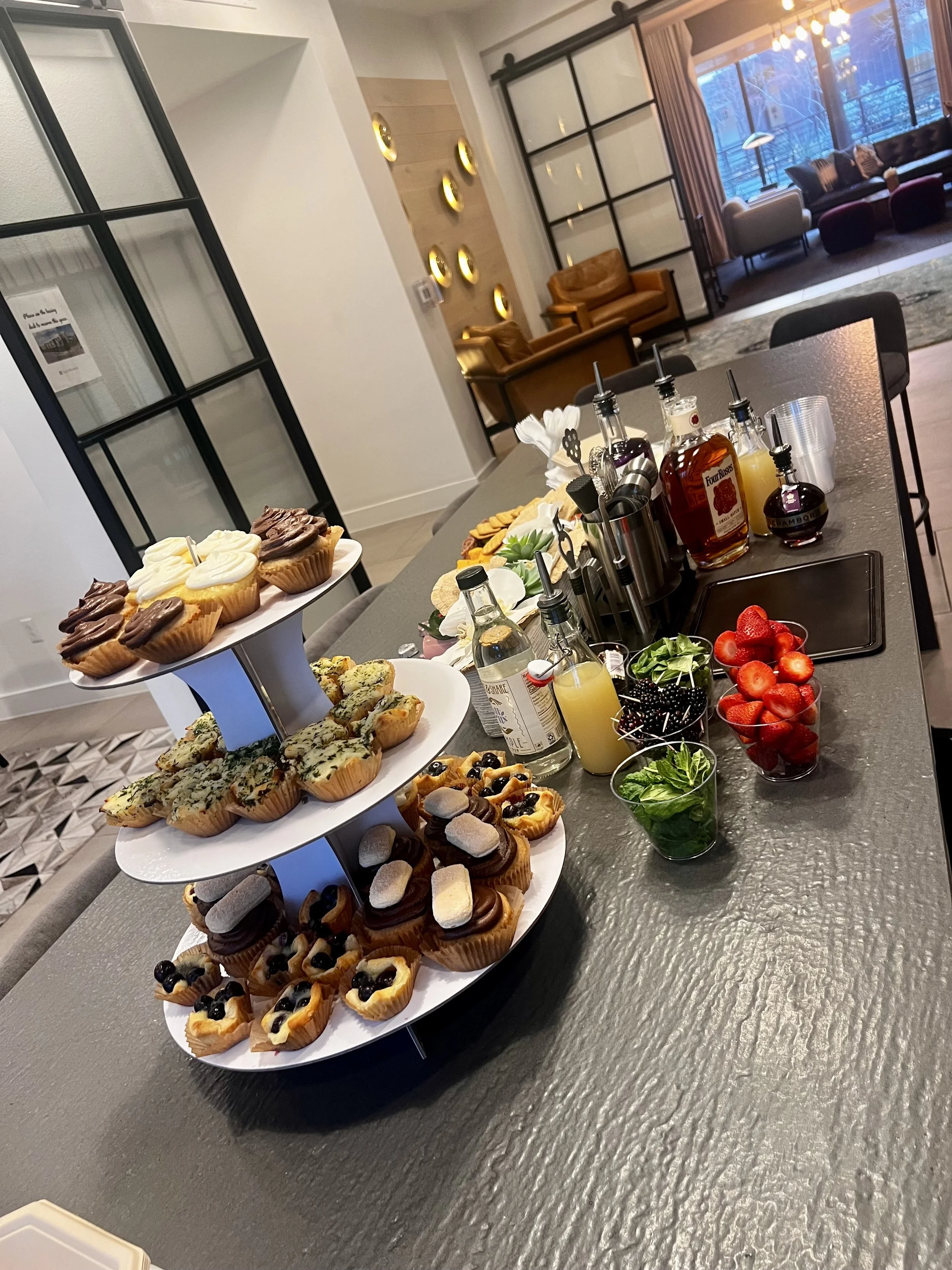 A table with a three-tiered stand holding cupcakes, muffins, and small tarts, surrounded by various bottles of drink mixers, fresh strawberries, blueberries, mint, and juice, set in a modern lounge with seating and large windows.
