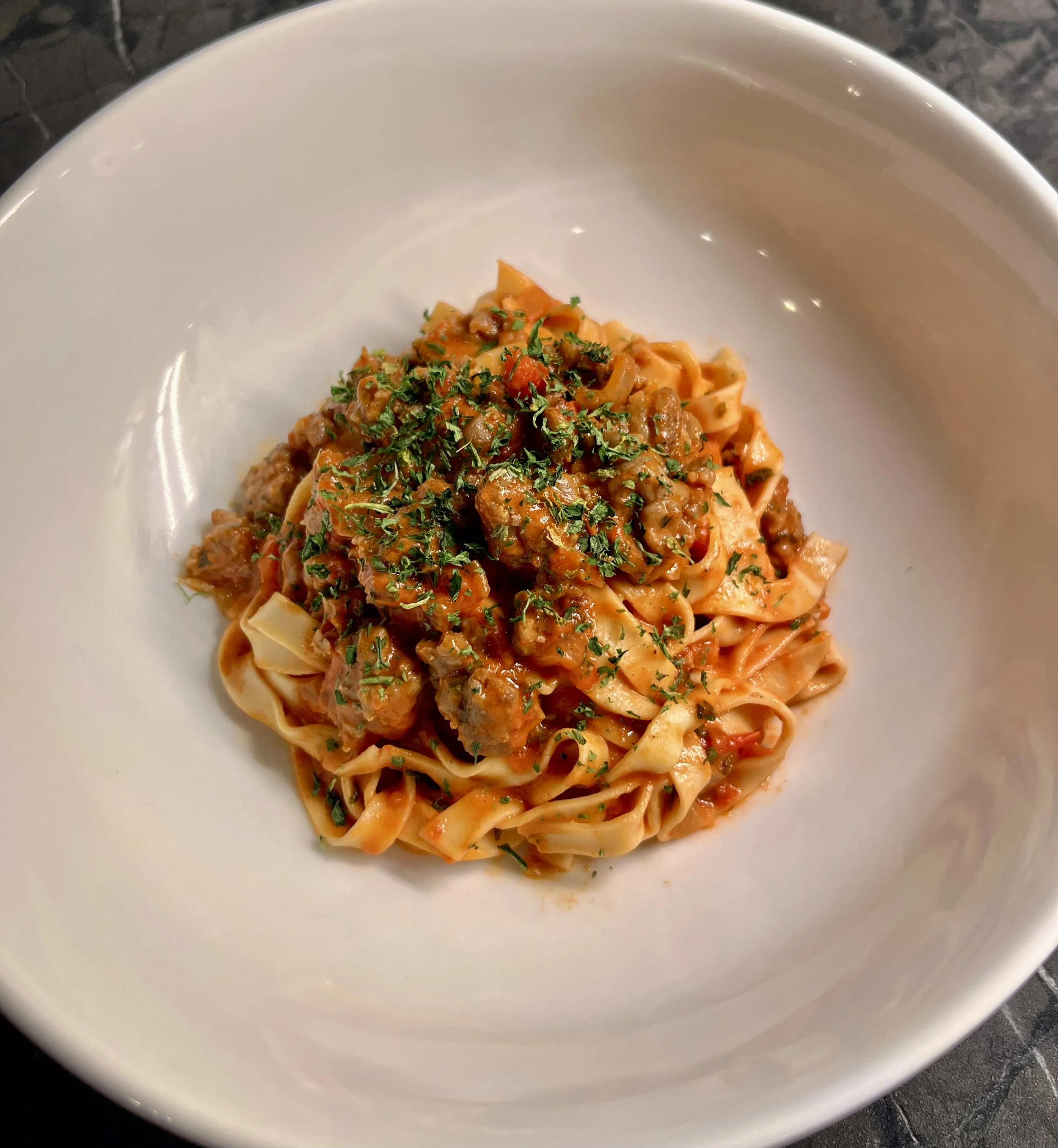 A white bowl of pasta with meat sauce and chopped parsley garnish.