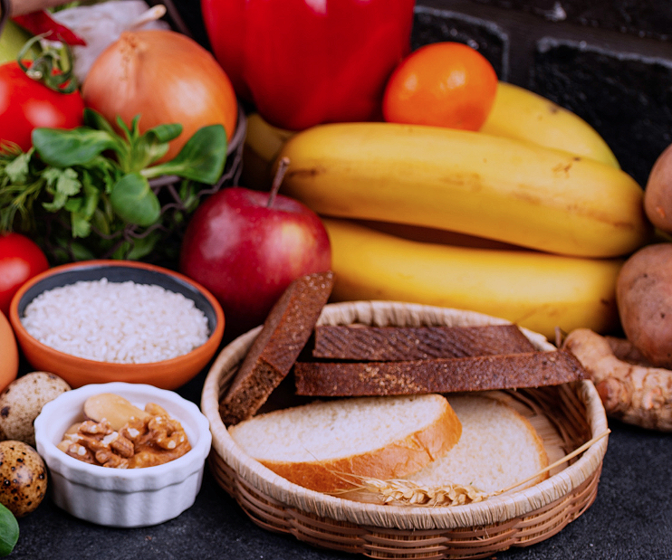 Assorted fresh fruits, vegetables, bread, and grains on a table, including bananas, apples, tomatoes, onions, and a basket of sliced bread and dark toast.