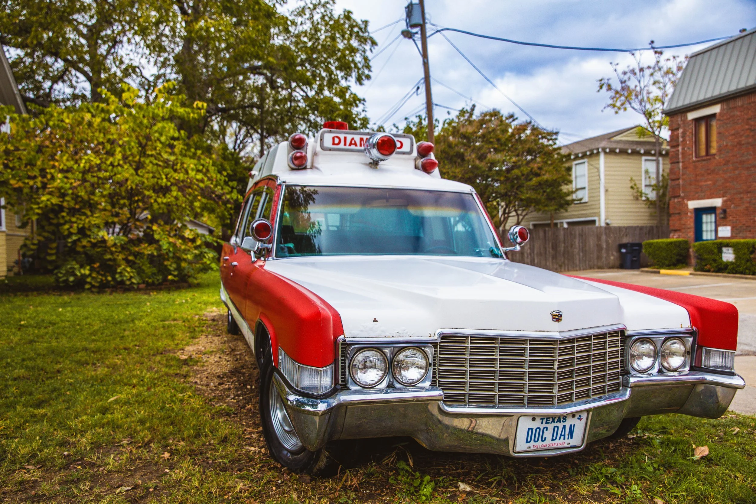 A vintage ambulance with a red and white color scheme, sirens, and a Texas license plate reading 'DOC DAN,' parked on grass in a suburban neighborhood.