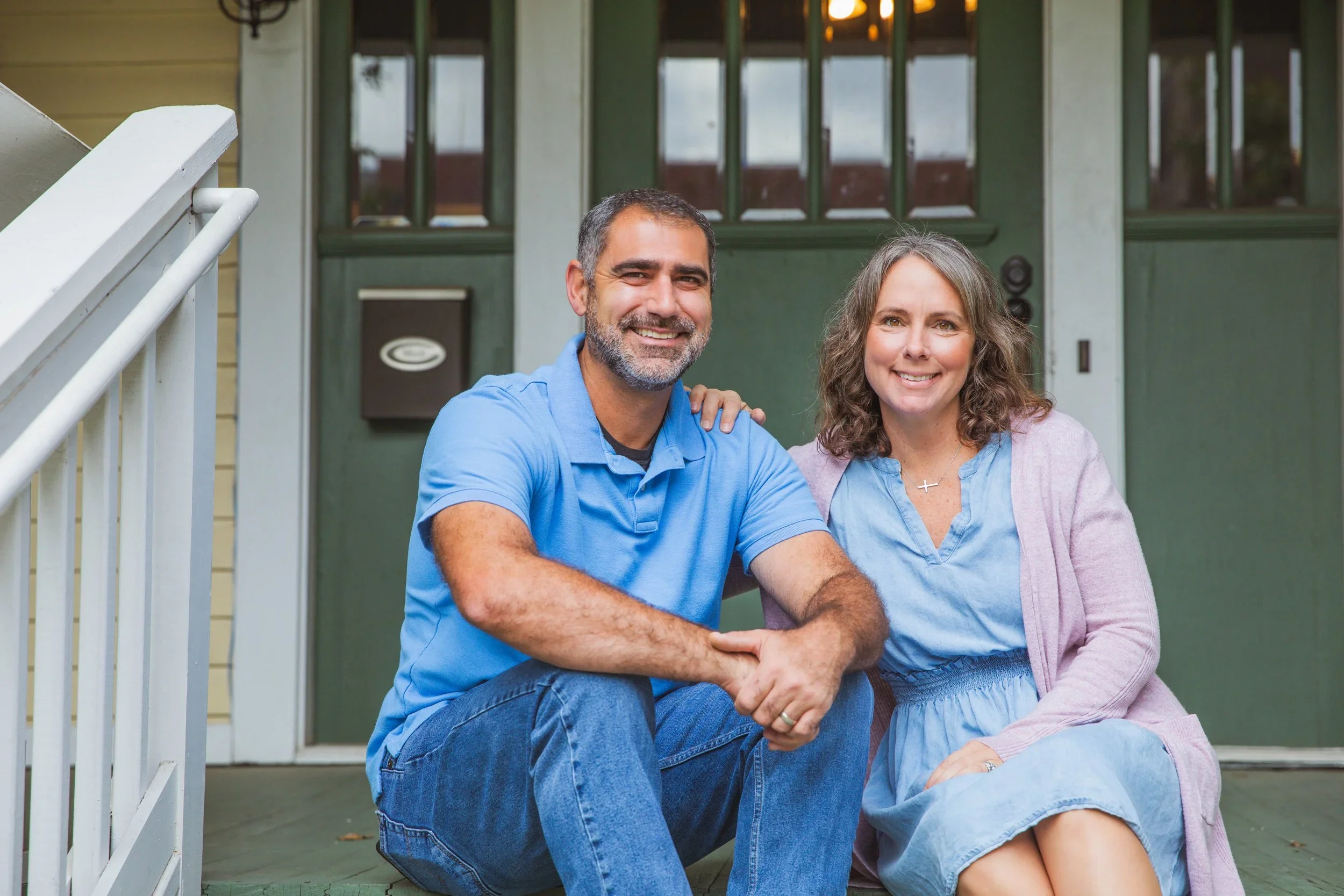 A smiling middle-aged couple sitting on the front porch of a house. The man is in a blue polo shirt and jeans, and the woman in a blue dress with a light pink cardigan. They are seated close together, with the man’s arm around the woman, and she has her hand on his shoulder.