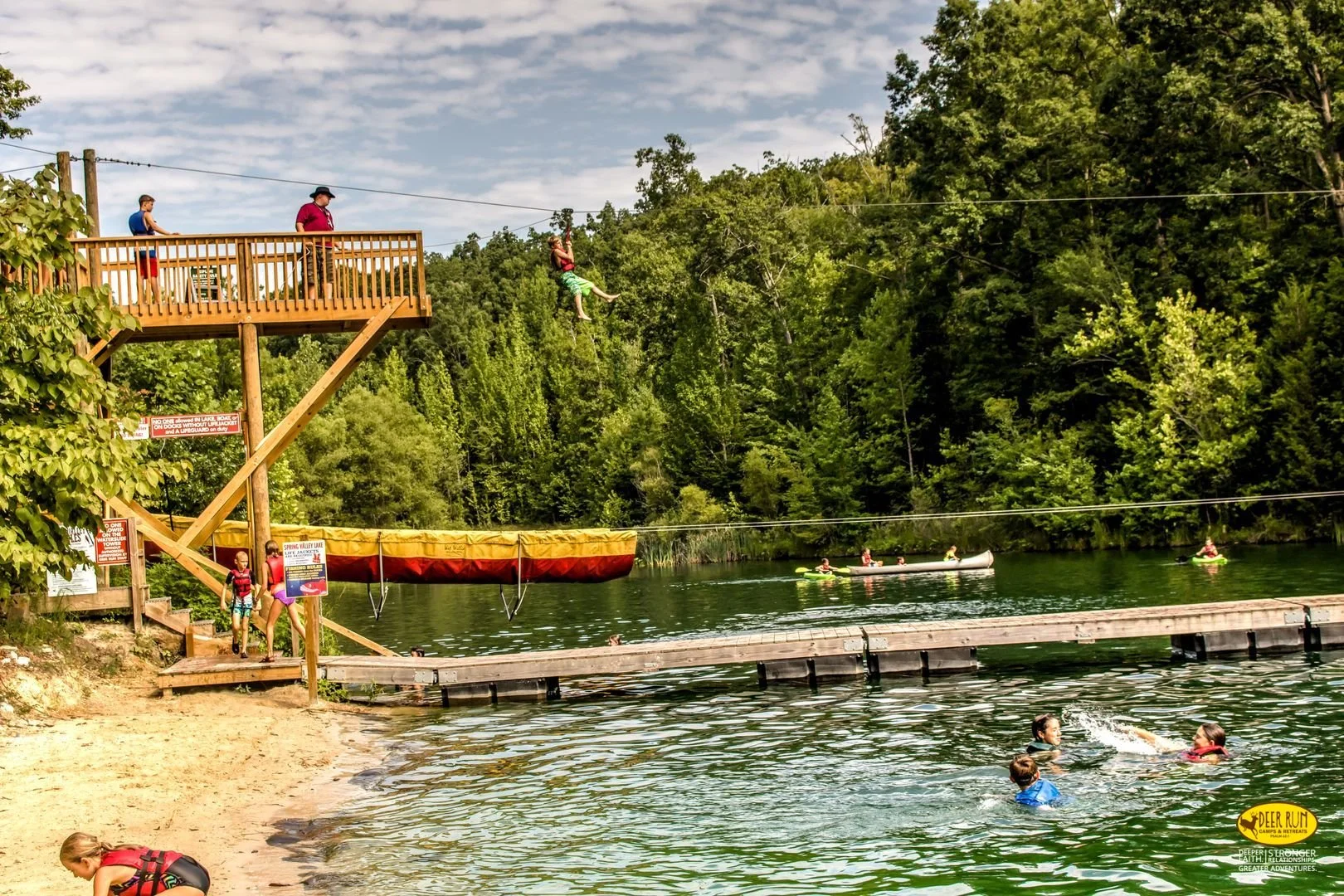 People enjoying a day at a lake with a zip line and kayaks, surrounded by green trees and a sandy shore.