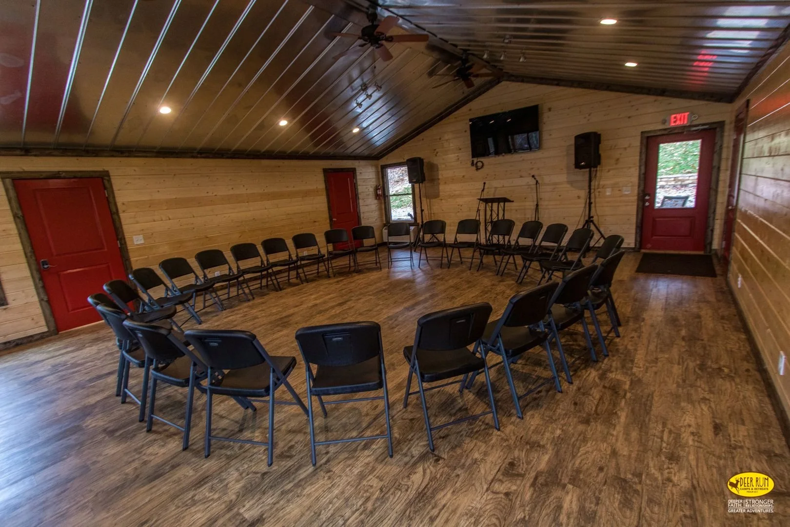 A round arrangement of black folding chairs inside a wooden cabin with wood paneling walls and ceiling, featuring a large flat-screen TV, microphones, and speakers, set up for a meeting or event.