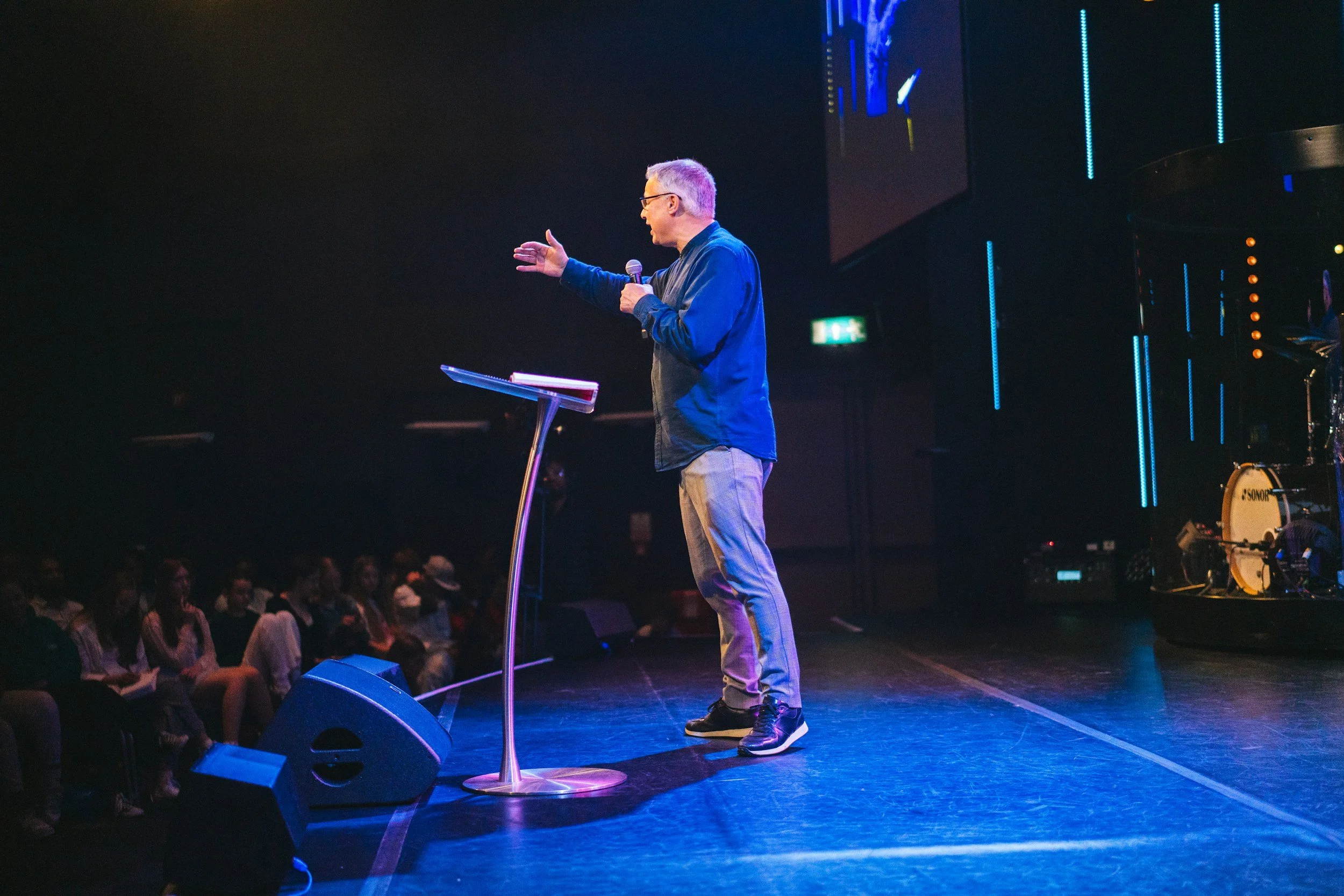 A man giving a speech or sermon on stage in a dark auditorium, holding a microphone and gesturing with his right hand, with an audience seated in front of him, stage lights and screen in the background.