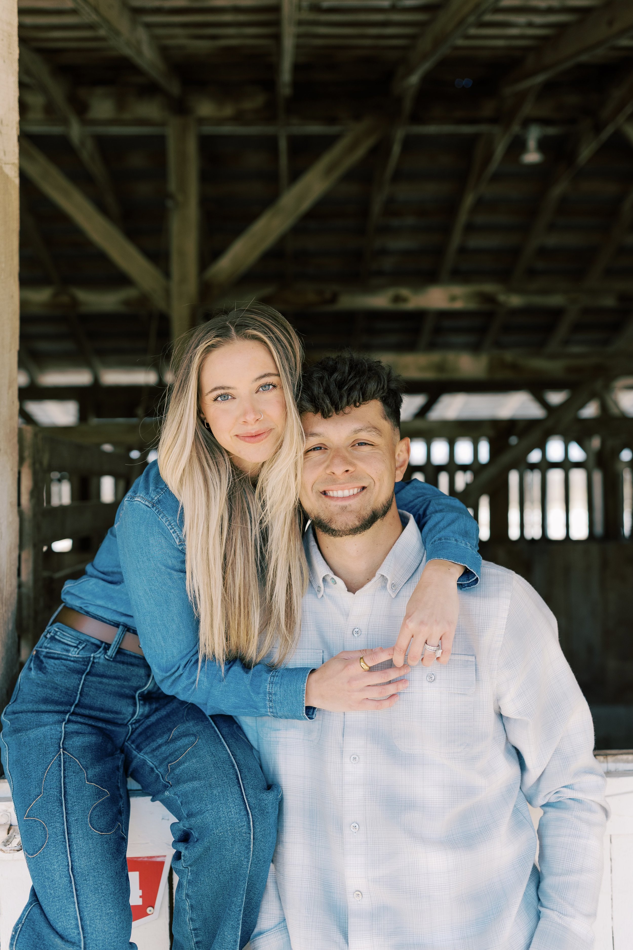 A young woman with long blonde hair in a blue denim shirt leaning on a young man with dark hair in a white shirt, both smiling, inside a rustic wooden barn.