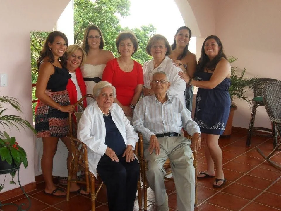 Group of nine women and elderly couple posing together indoors with a window and greenery outside.