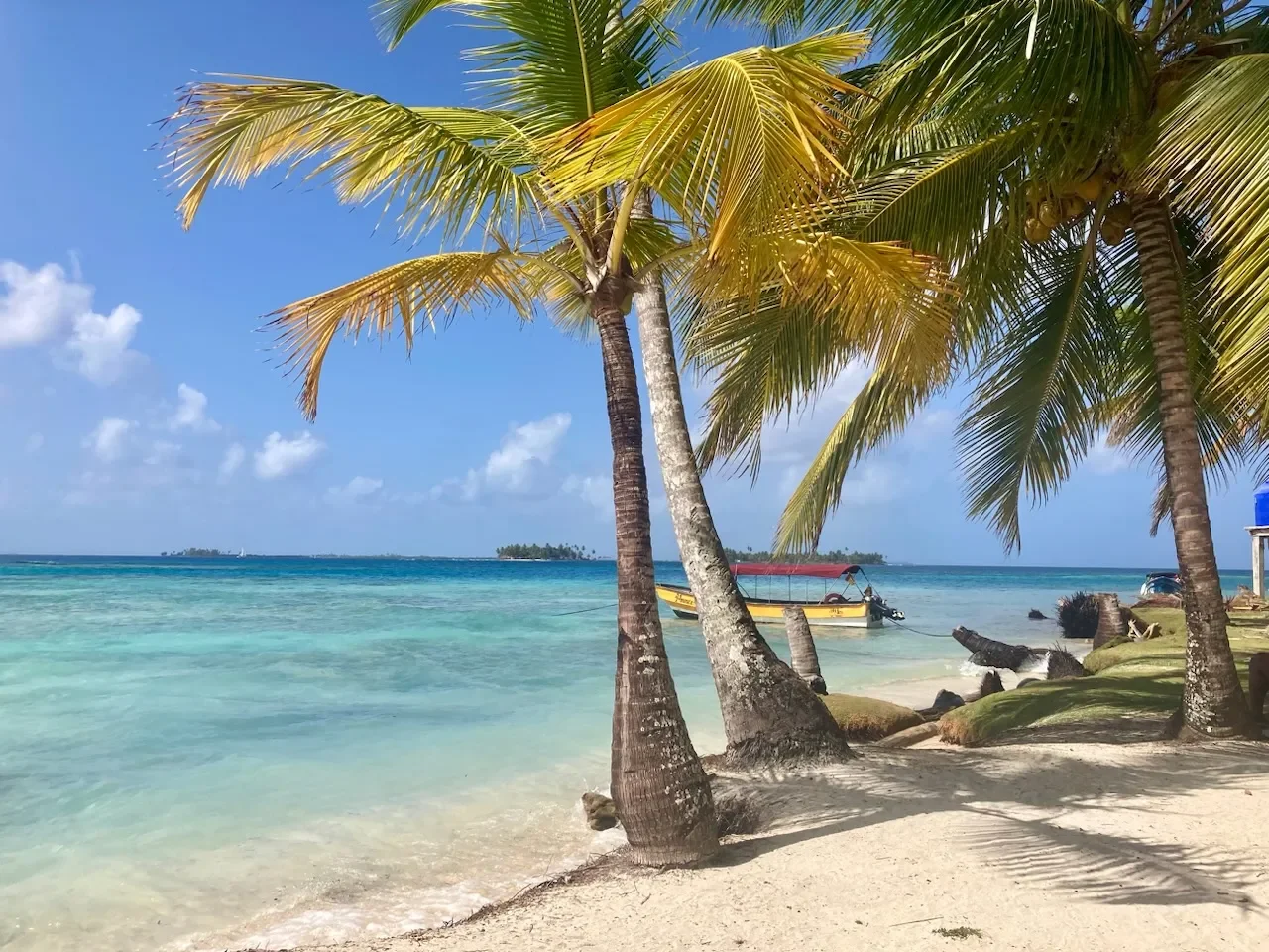 Tropical beach scene with palm trees, clear turquoise water, a yellow boat anchored near the shore, and a sunny blue sky.