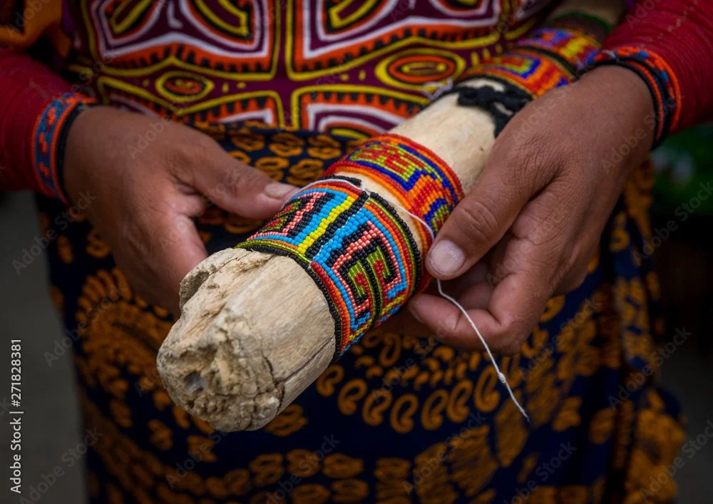Close-up of a person's hands holding a decorated wooden stick with colorful beadwork, wearing traditional clothing and bracelets.