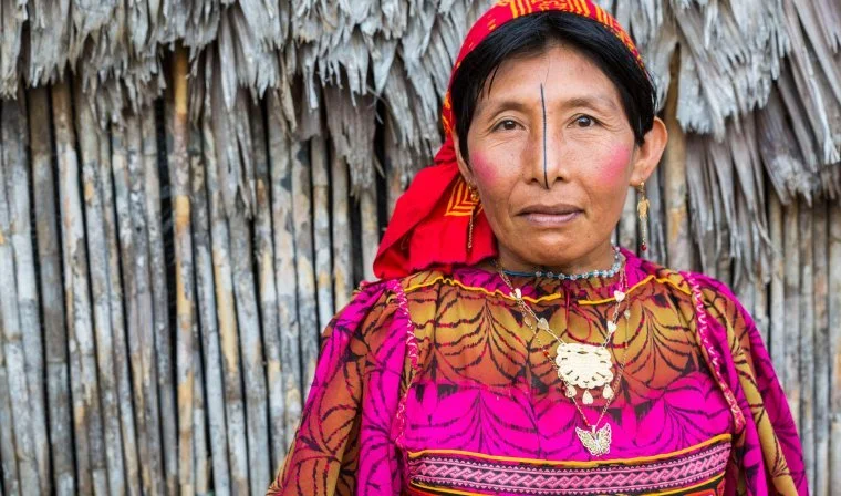 A woman dressed in traditional colorful clothing standing in front of a thatched wall.
