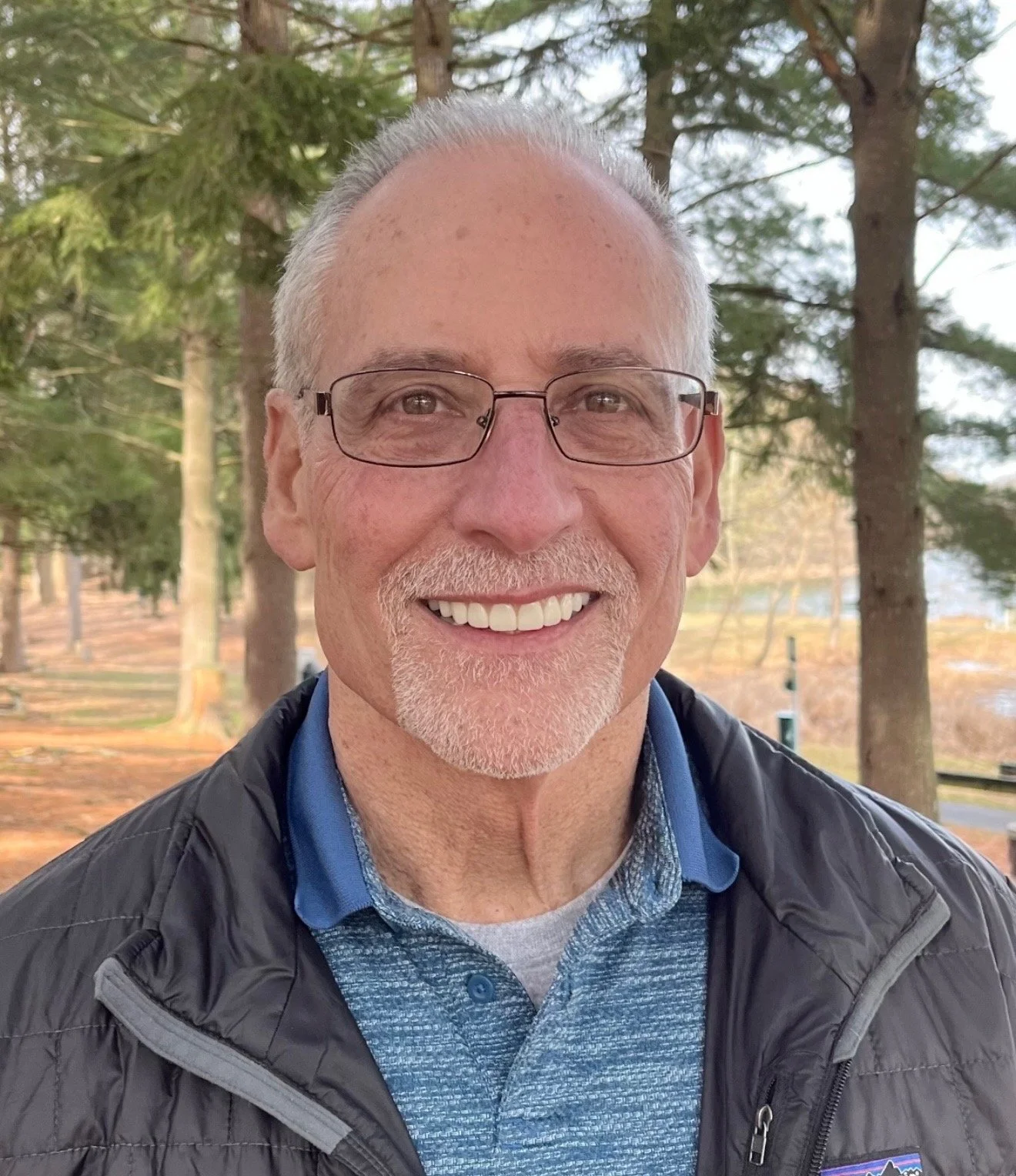 A smiling older man with glasses, a white beard, and gray hair, wearing a blue collared shirt and a black jacket, standing outdoors among trees.