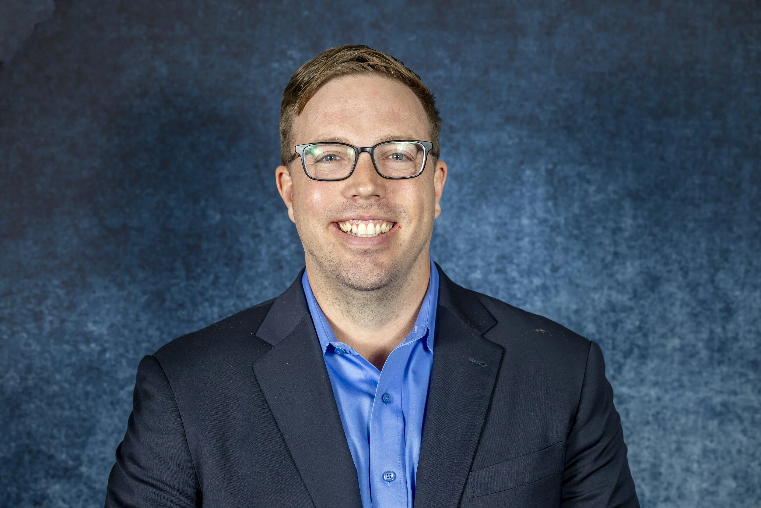 A man with glasses, smiling, wearing a blue shirt and a dark blazer, against a textured blue background.