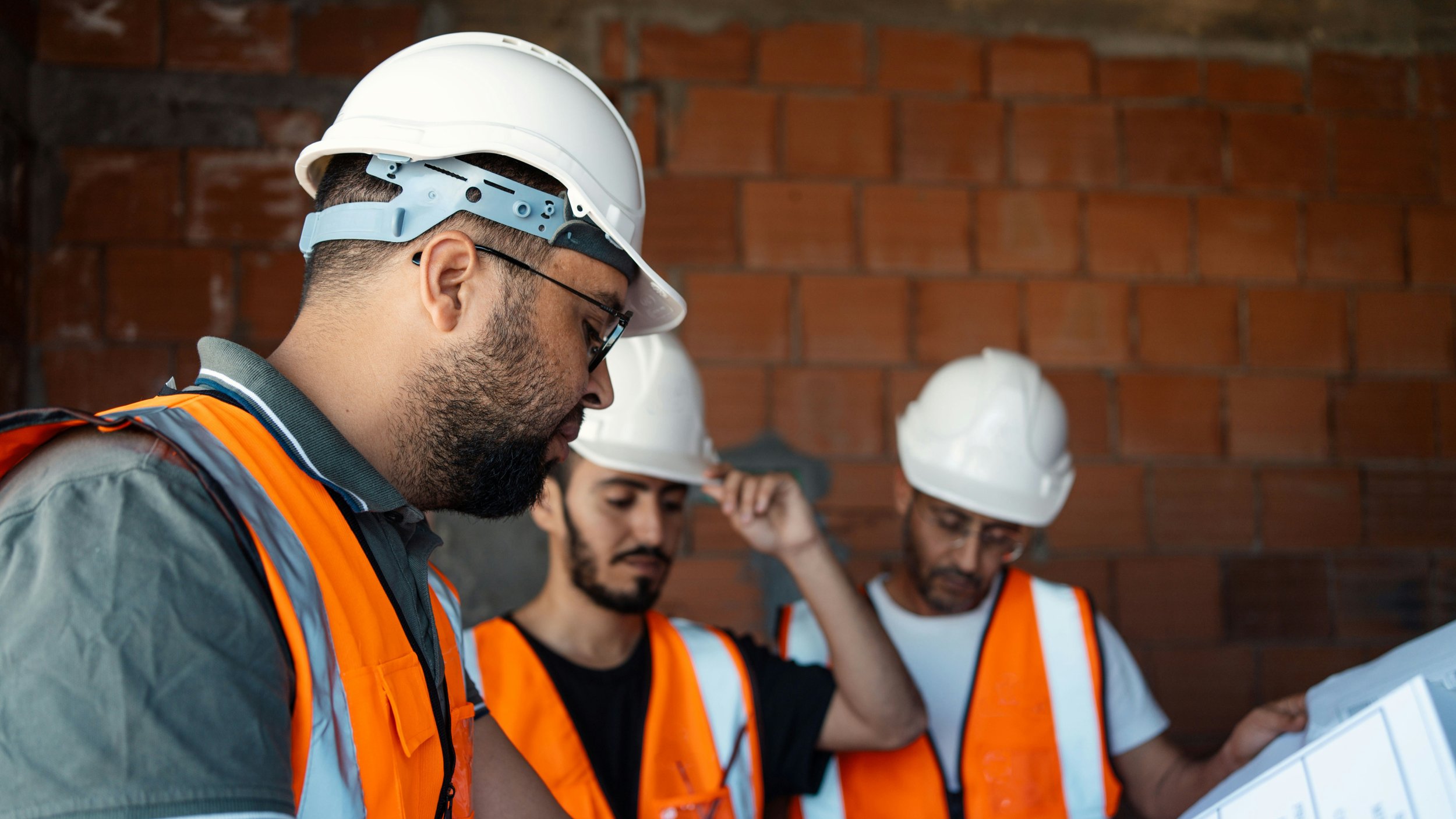 Construction workers wearing white safety helmets and orange safety vests examining blueprints against a brick wall background.