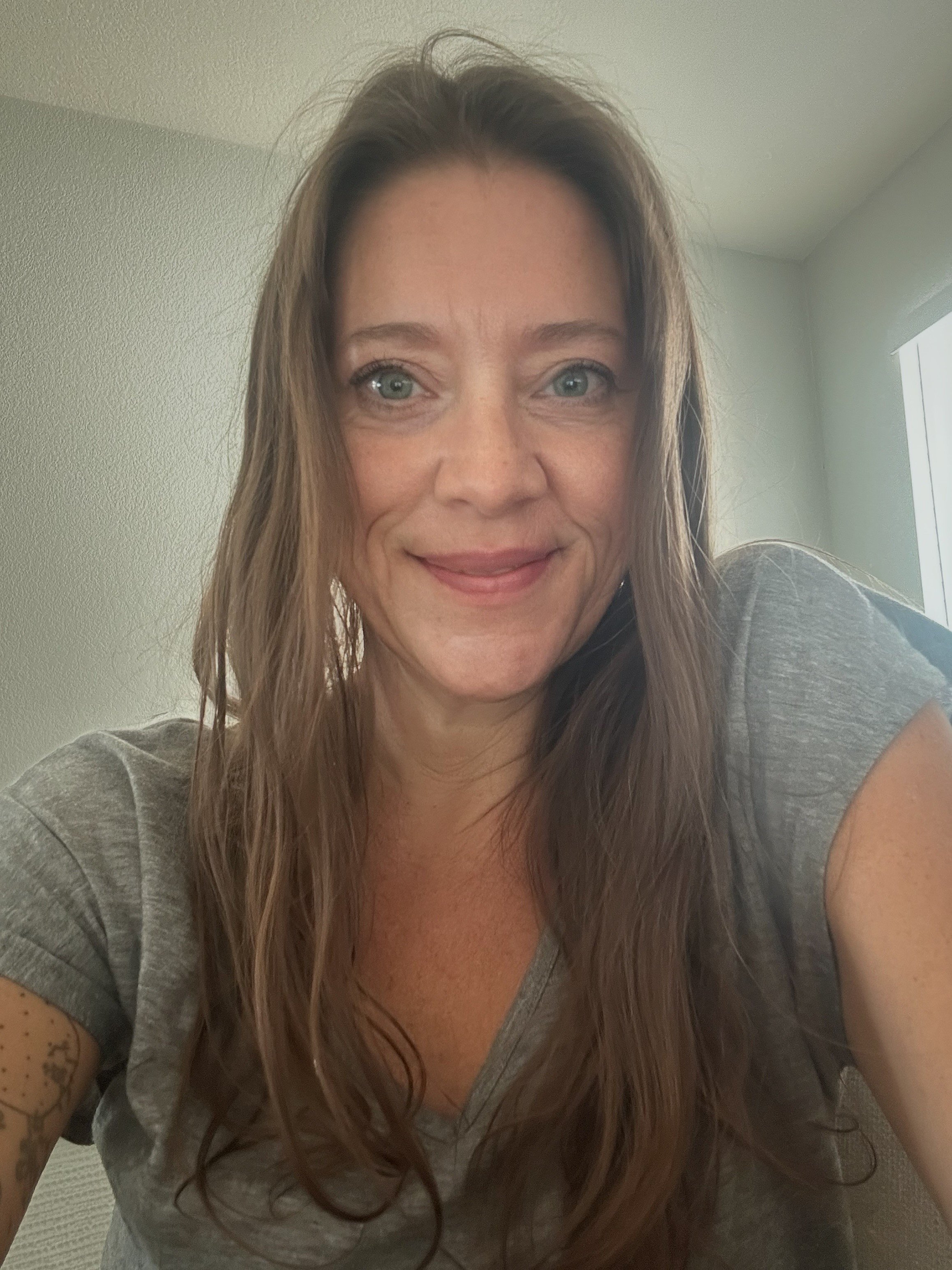 Close-up portrait of a smiling woman with long brown hair, wearing a gray t-shirt, in a room with light-colored walls and a window.