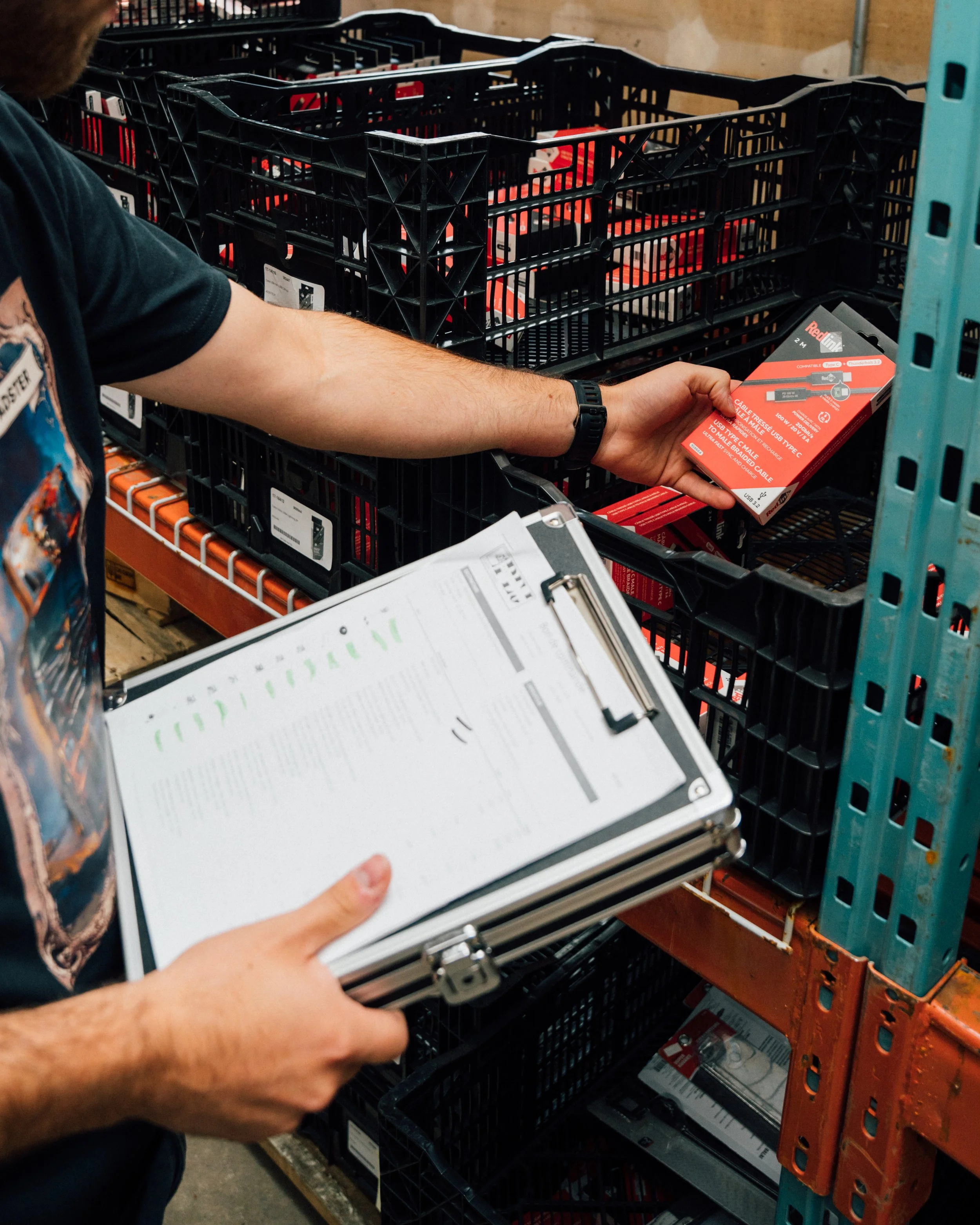 Un homme tenant une boîte rouge contenant un câble USB dans un entrepôt ou une section de magasin, avec des étagères métalliques remplies de produits similaires.