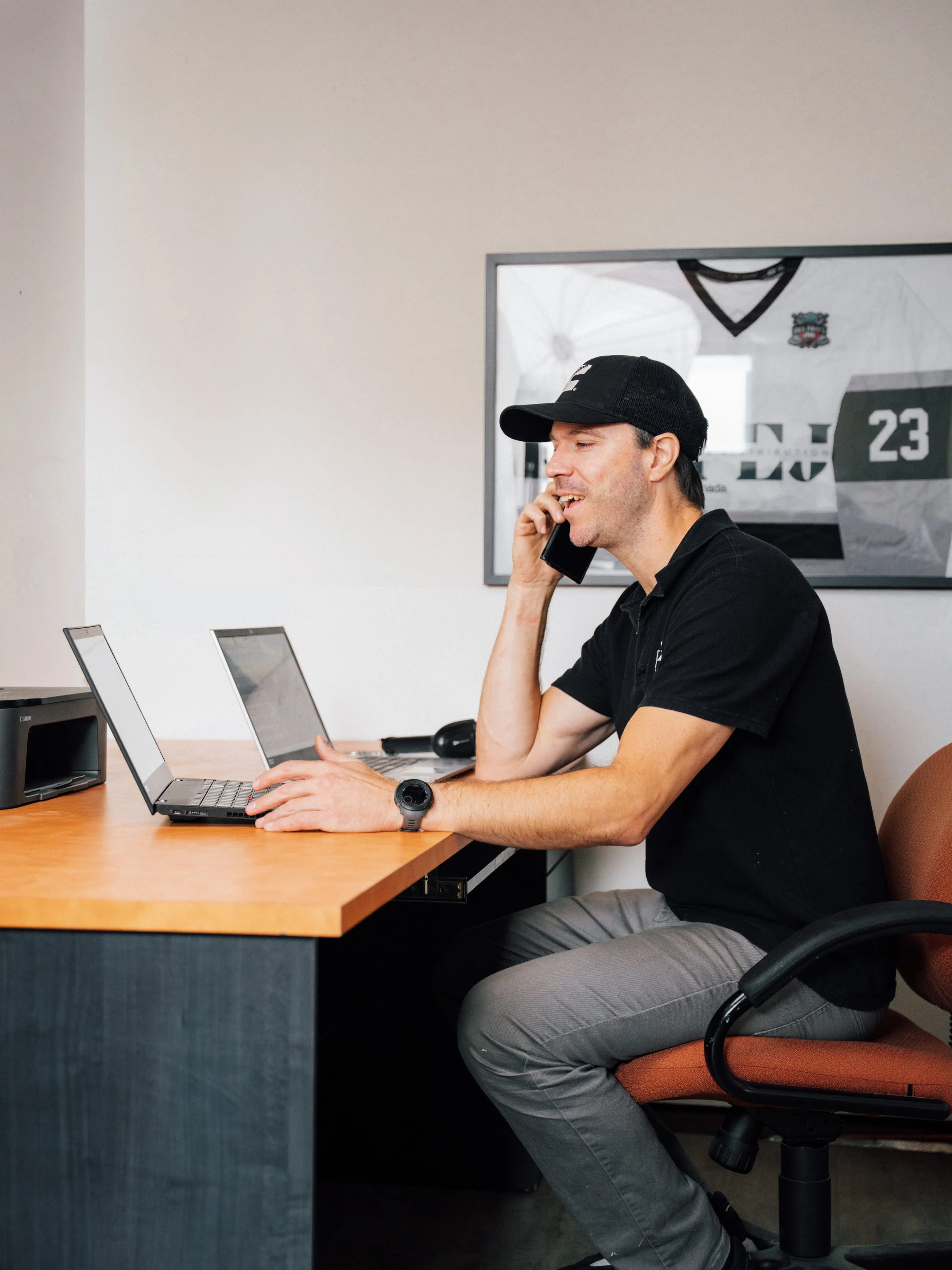Un homme en polo noir, portant une casquette noire, assis à un bureau en train de parler au téléphone et utilisant un ordinateur portable.