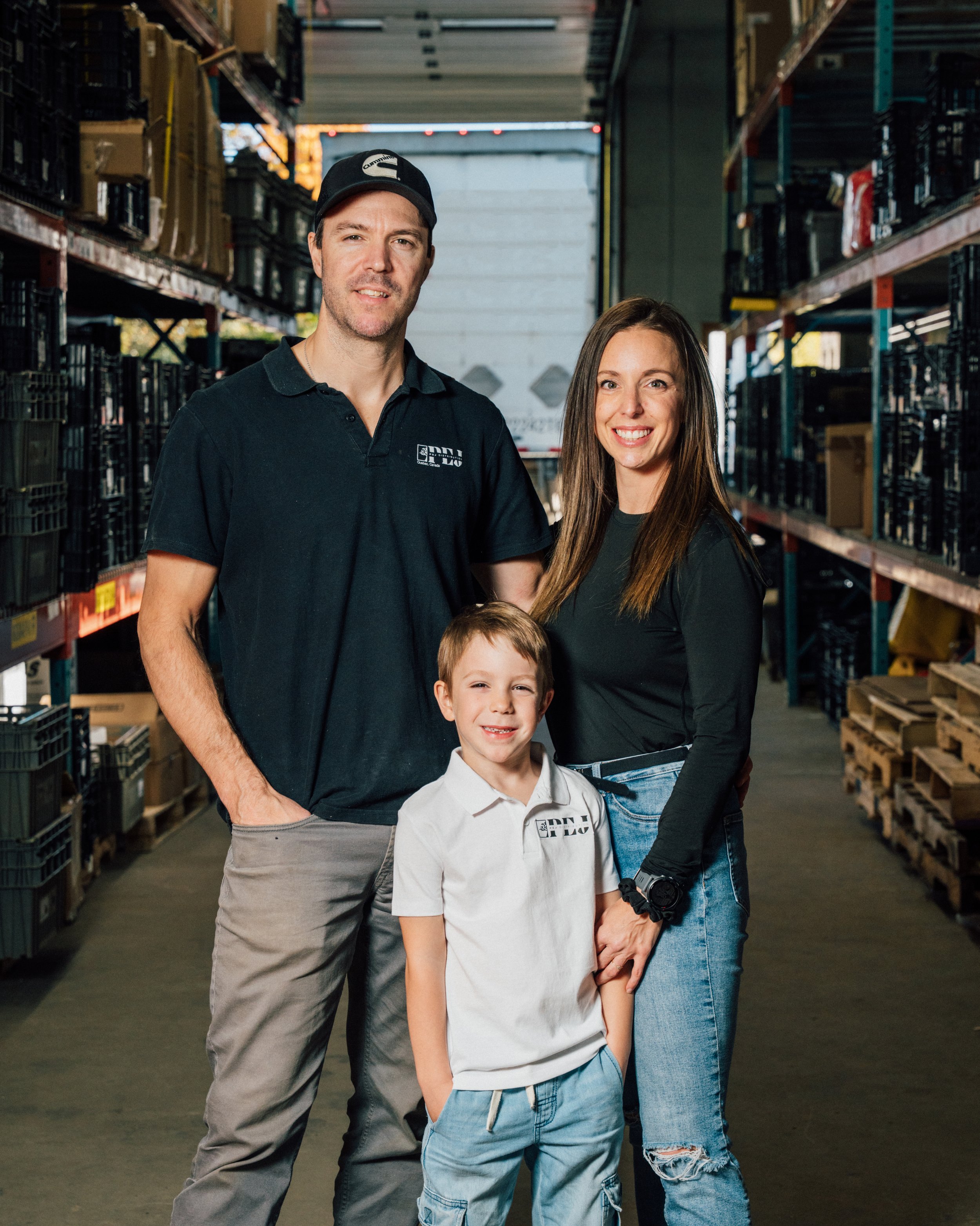 Une famille avec un homme, une femme et un garçon debout dans un entrepôt ou un stockage. Ils sourient et portent des vêtements décontractés. Les étagères derrière eux sont remplies de caisses et de boîtes.