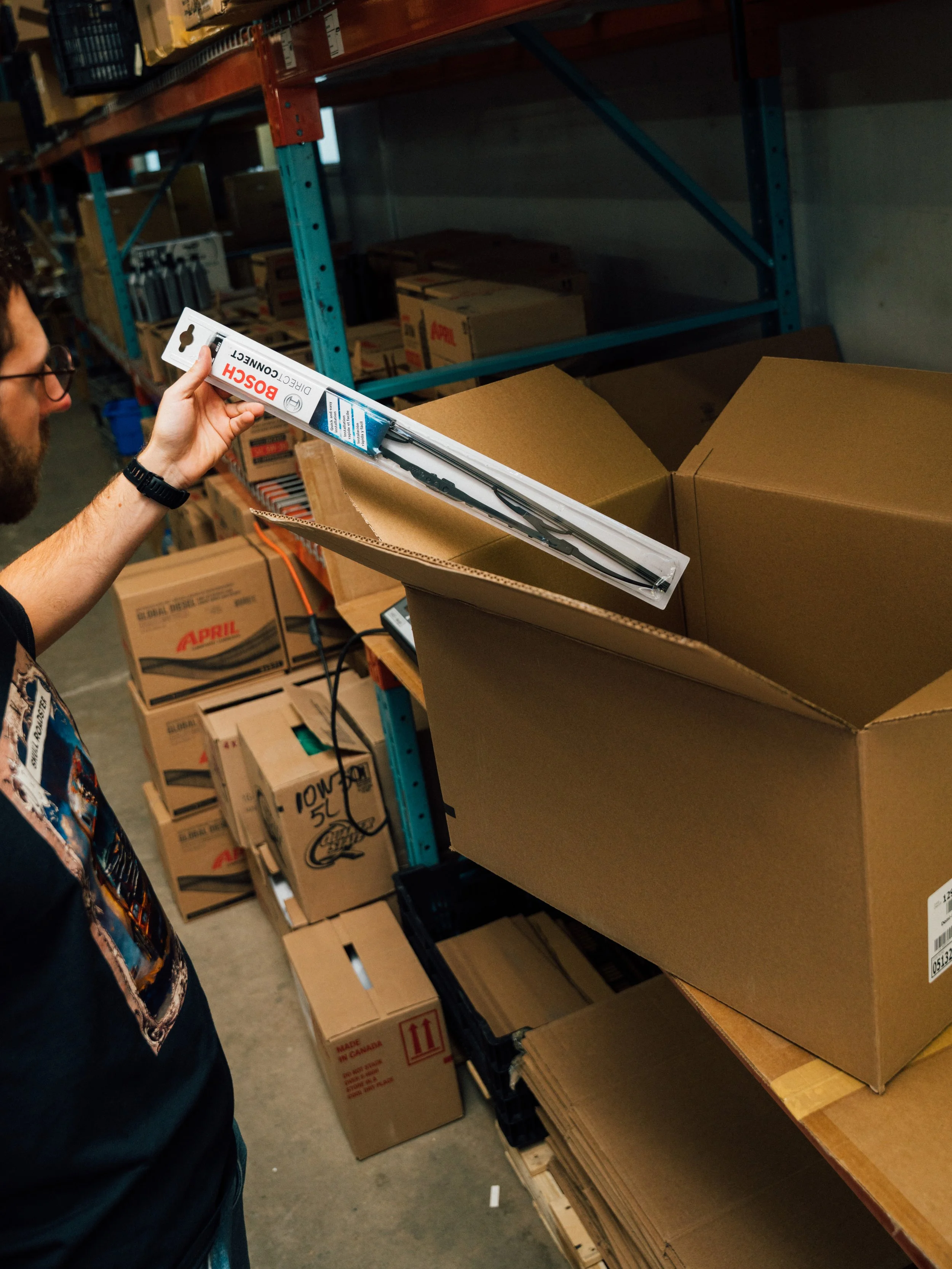 Une personne tient une brosse d’essuie-glace Bosch dans un entrepôt de stockage, regardant une grande boîte en carton ouverte sur une palette. L'entrepôt est rempli de cartons sur des étagères métalliques.