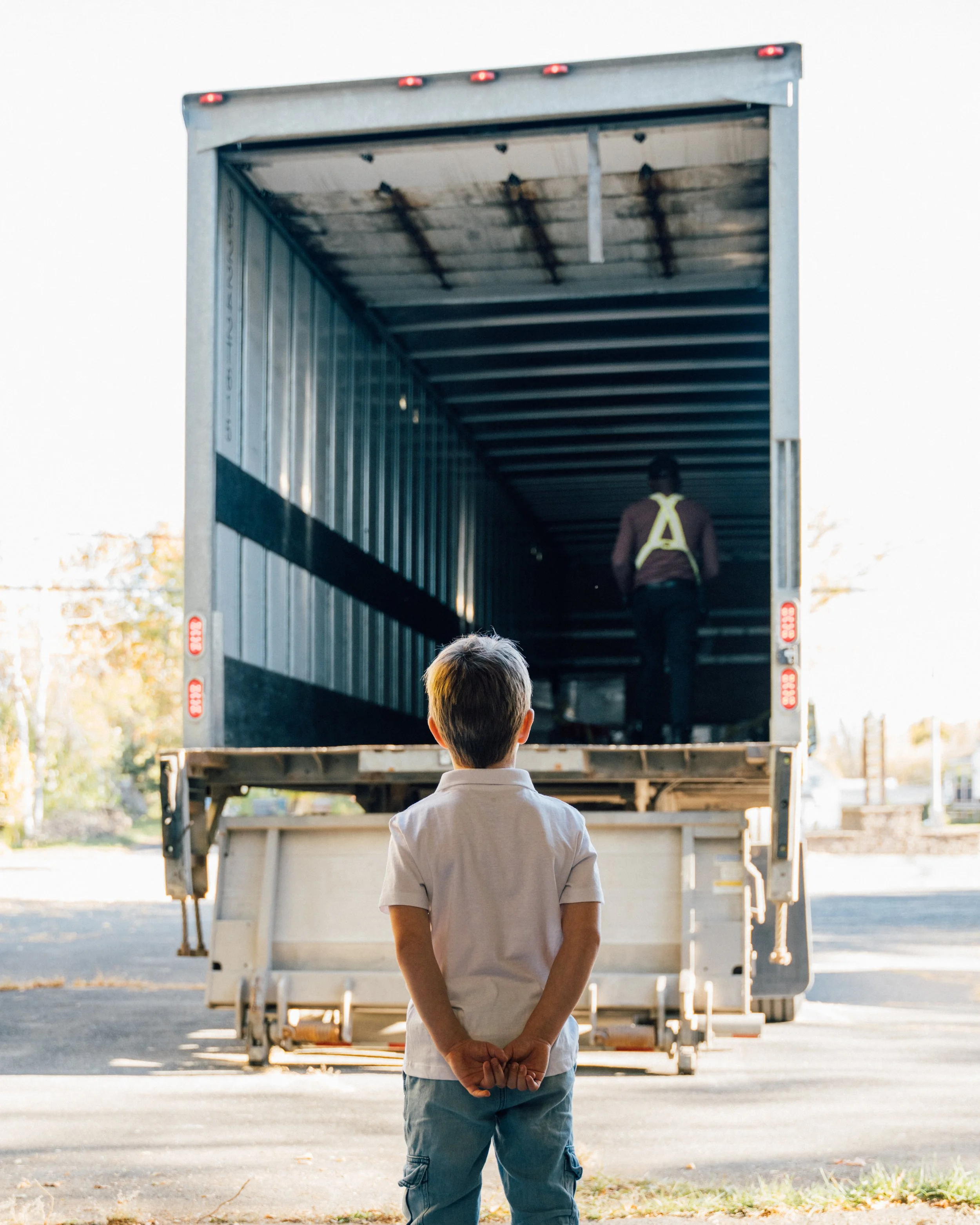 Un garçon regarde un camion de déménagement avec un adulte à l'intérieur, stationné en extérieur, avec des arbres en arrière-plan.
