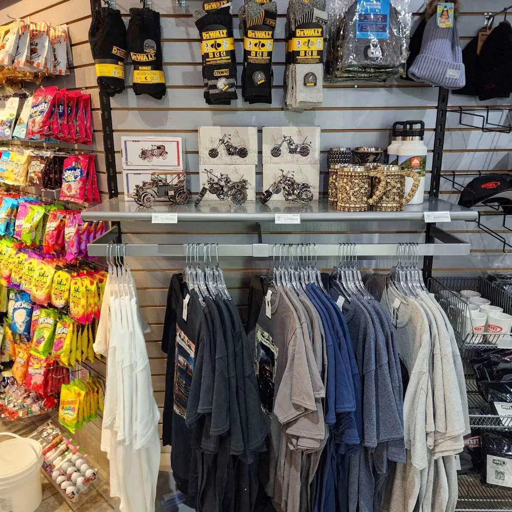 Vêtements, accessoires et décorations dans un magasin, avec des T-shirts, des mugs, des boîtes en bois, des gants, un chapeau, un sac à dos, et des articles pour l'extérieur.