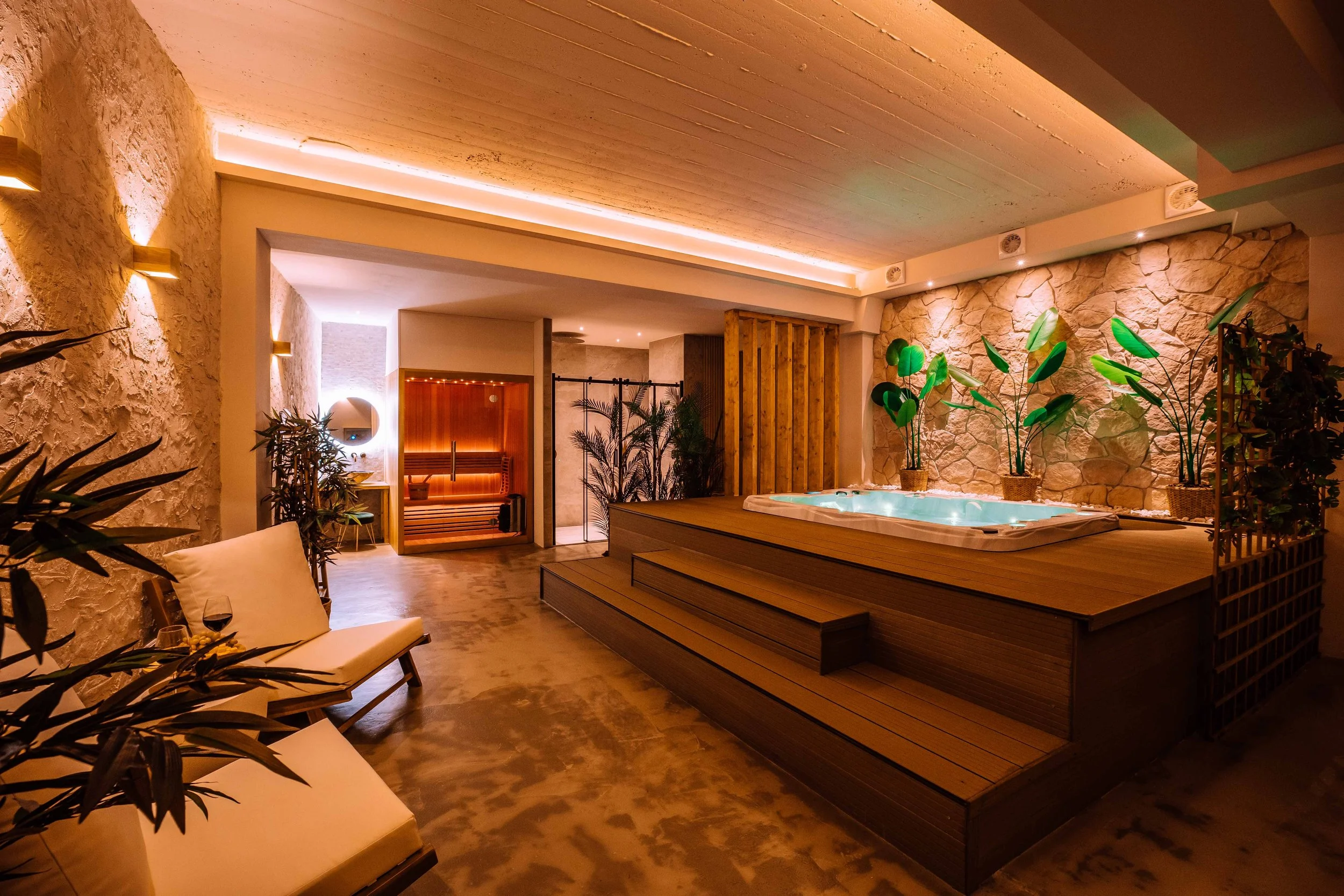 Indoor spa room with a hot tub on a raised wooden platform, surrounded by lush green plants and warm ambient lighting, with relaxed seating areas and sauna in the background.