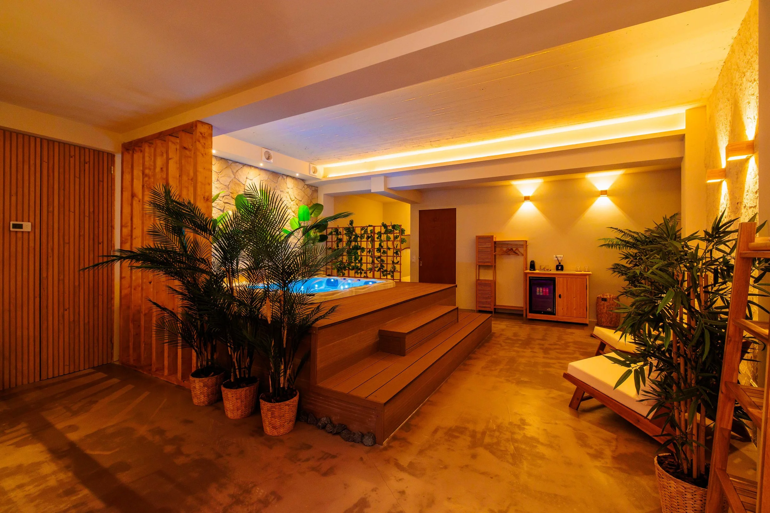 Indoor spa room with jacuzzi on wooden platform, green plants, warm yellow lighting, and wooden furniture.