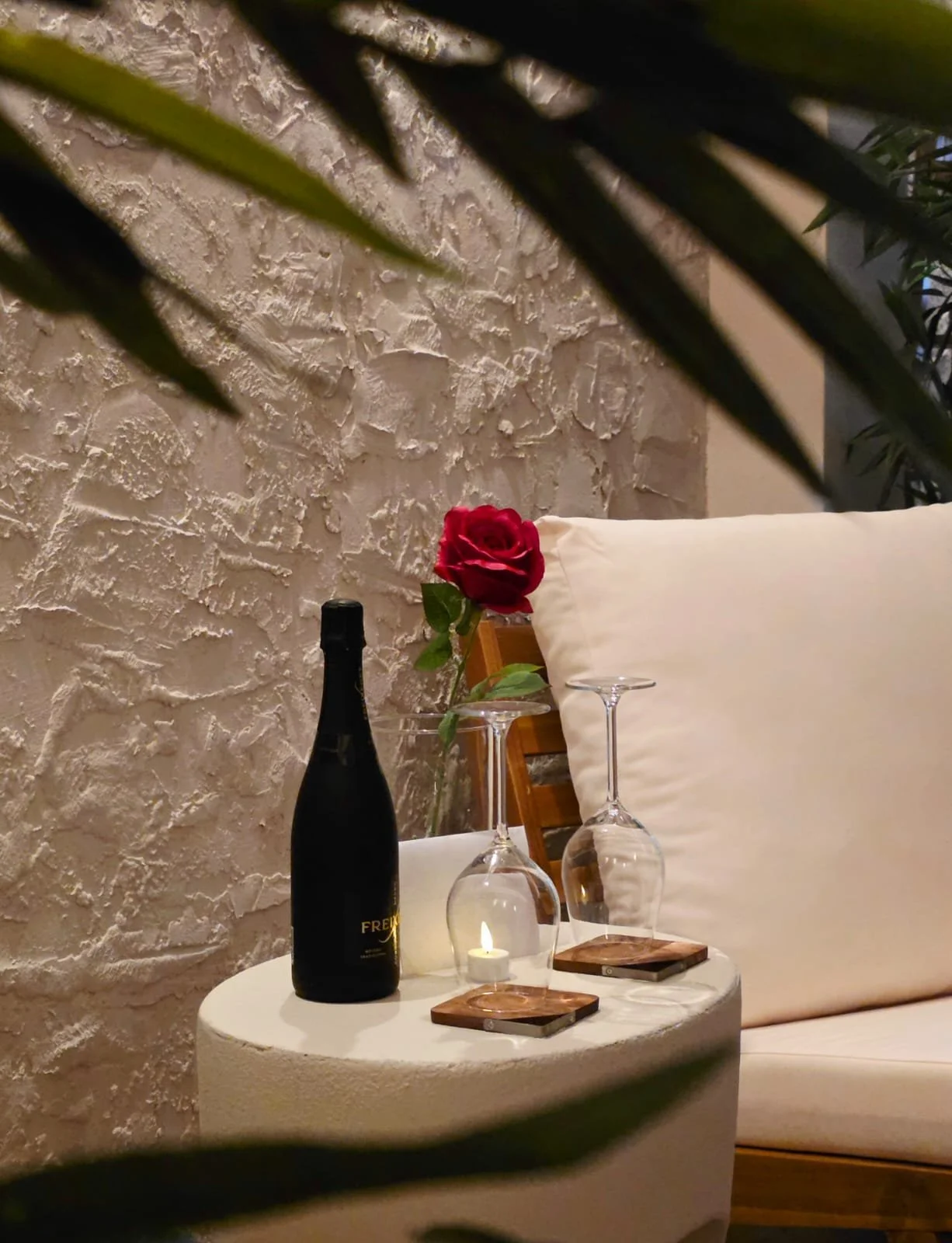 A cozy sitting area with a small white table holding a wine bottle, two upside-down wine glasses, a lit candle, and a single red rose in a glass vase for romantic settings, set against a textured white stone wall and a cushioned bench.