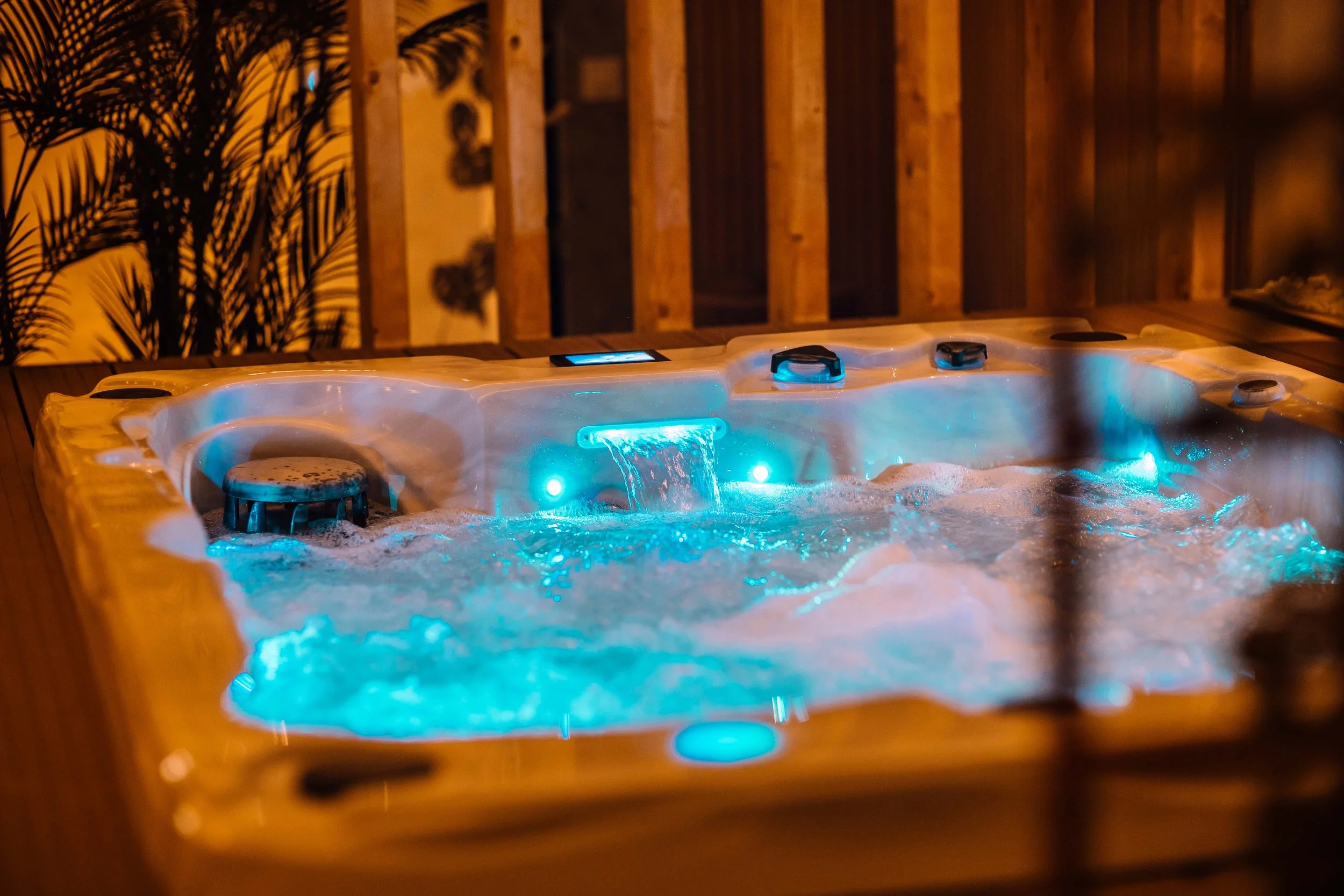 A jacuzzi hot tub with blue bubbling water, with an illuminated waterfall feature, surrounded by a wooden deck and a background of wooden walls and plants in a cozy outdoor setting at night.